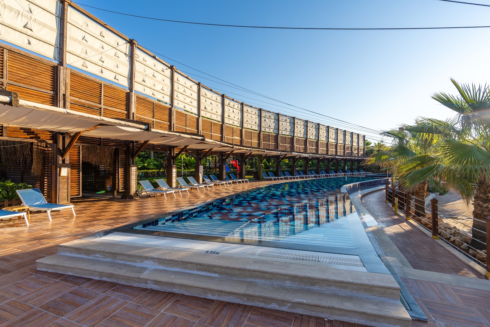A long, outdoor swimming pool featuring a unique architectural design with high, decorative wooden walls providing significant visual seclusion on one side, surrounded by lounging areas.