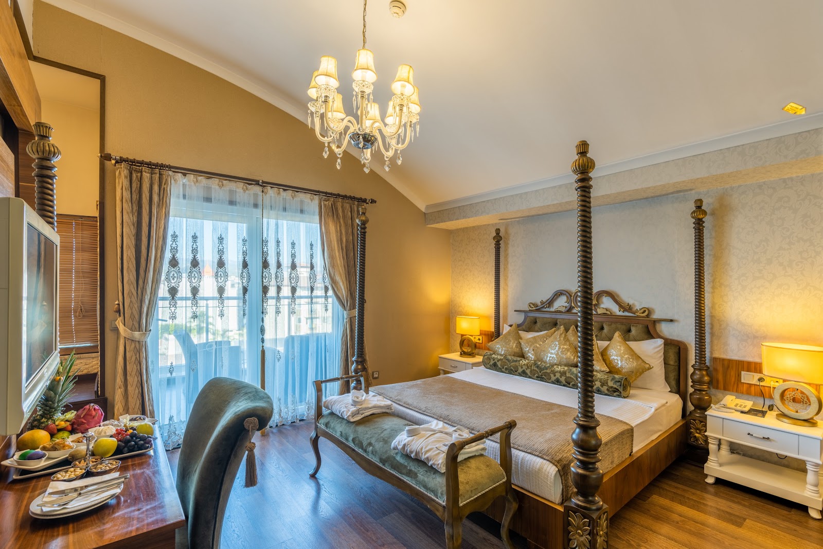 An elegantly furnished hotel room with a luxurious four-poster king-sized bed, ornate chandelier, a comfortable chaise lounge, and a private balcony accessible through large glass doors.