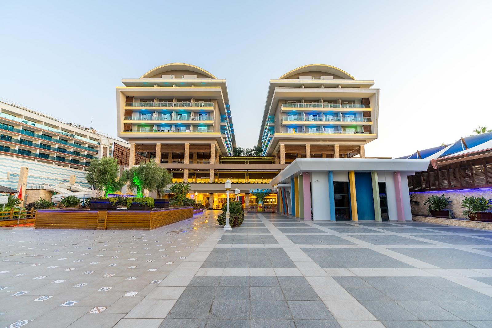 The striking modern facade of the Adenya Hotel & Resort, featuring multi-story buildings with colorful illuminated balconies and a spacious entrance area.
