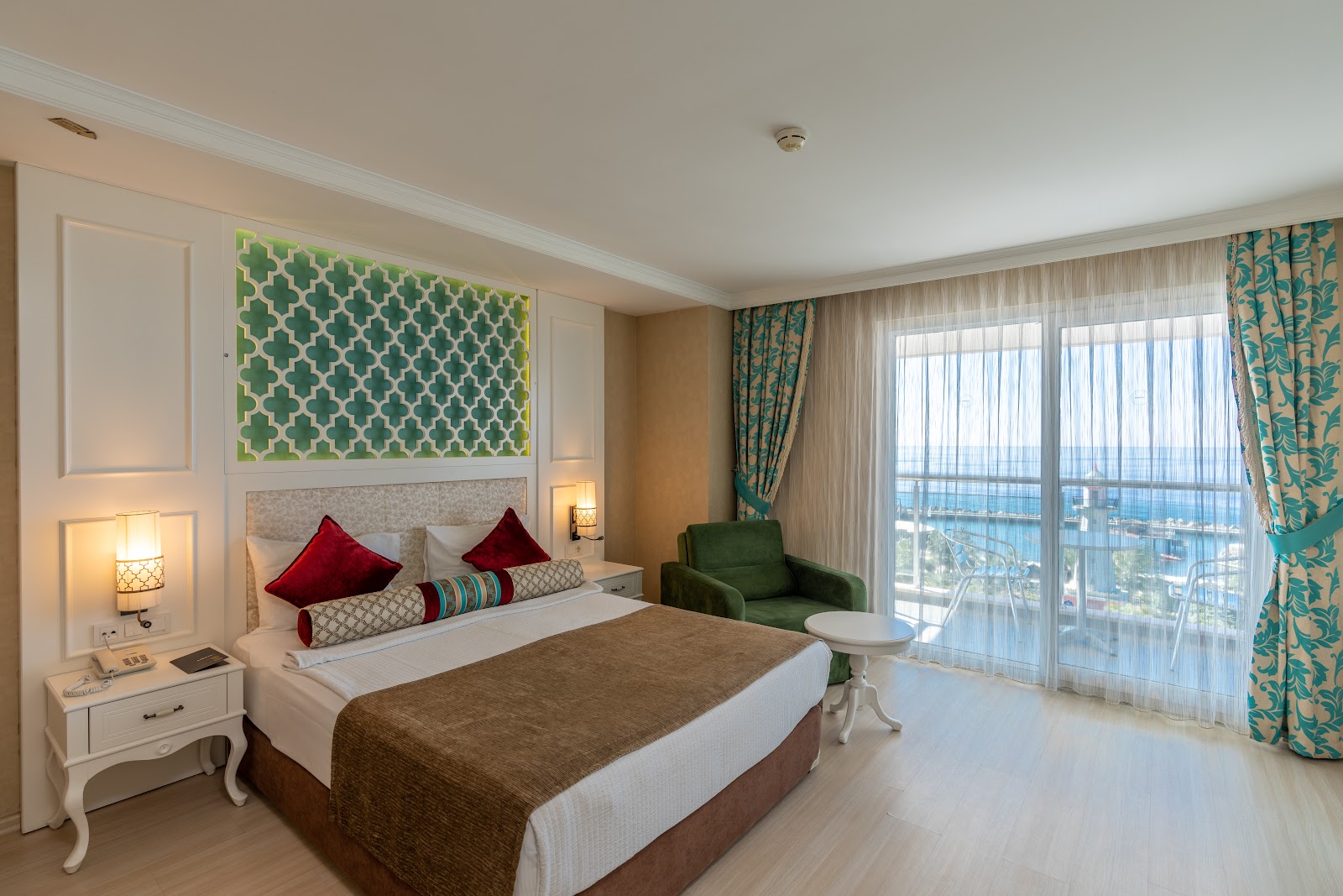 A bright and modern hotel room with a king-sized bed, a decorative patterned headboard, a green armchair, and a private balcony offering a beautiful sea view.