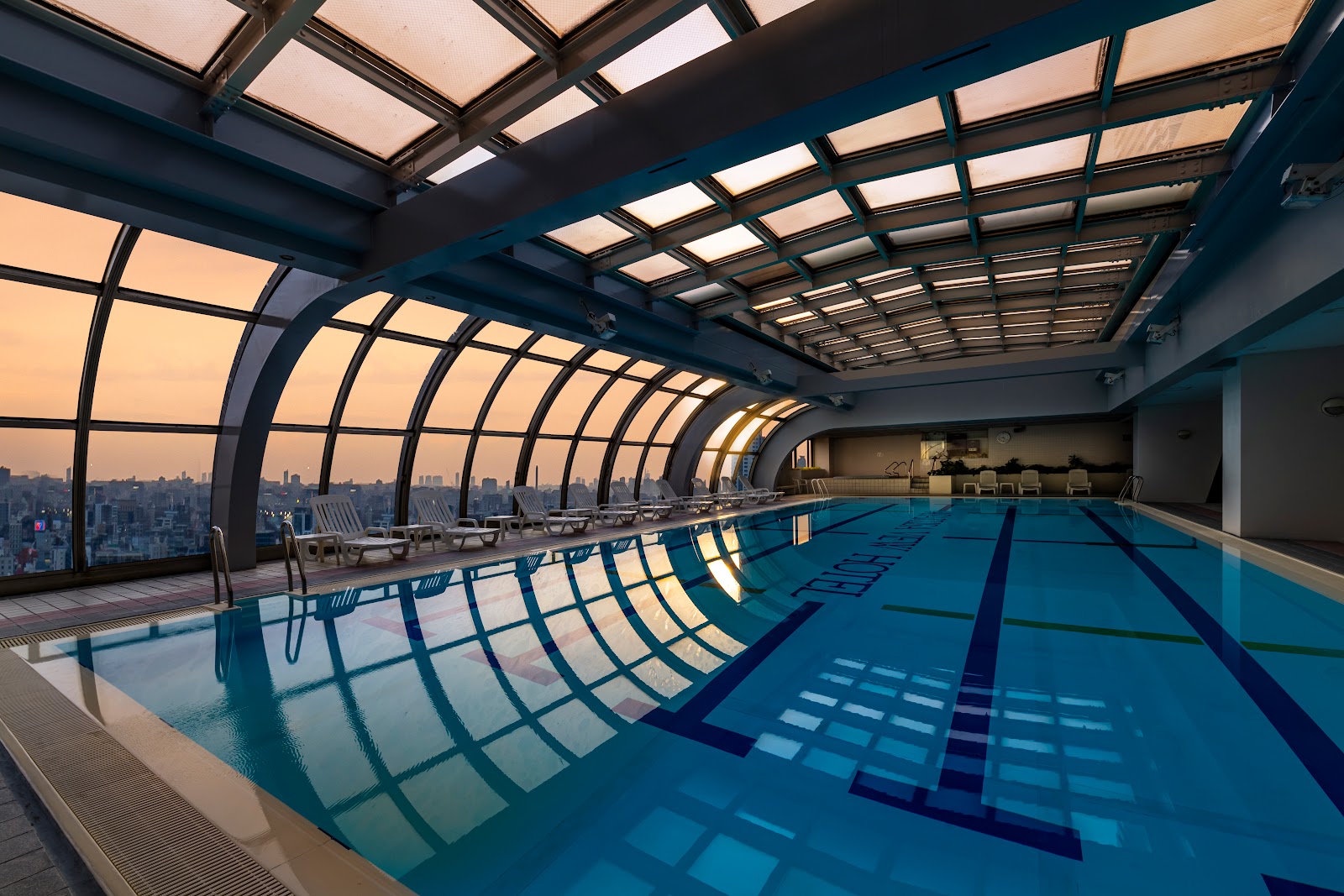 An empty indoor swimming pool with a unique arched glass roof and large windows offering panoramic city views.