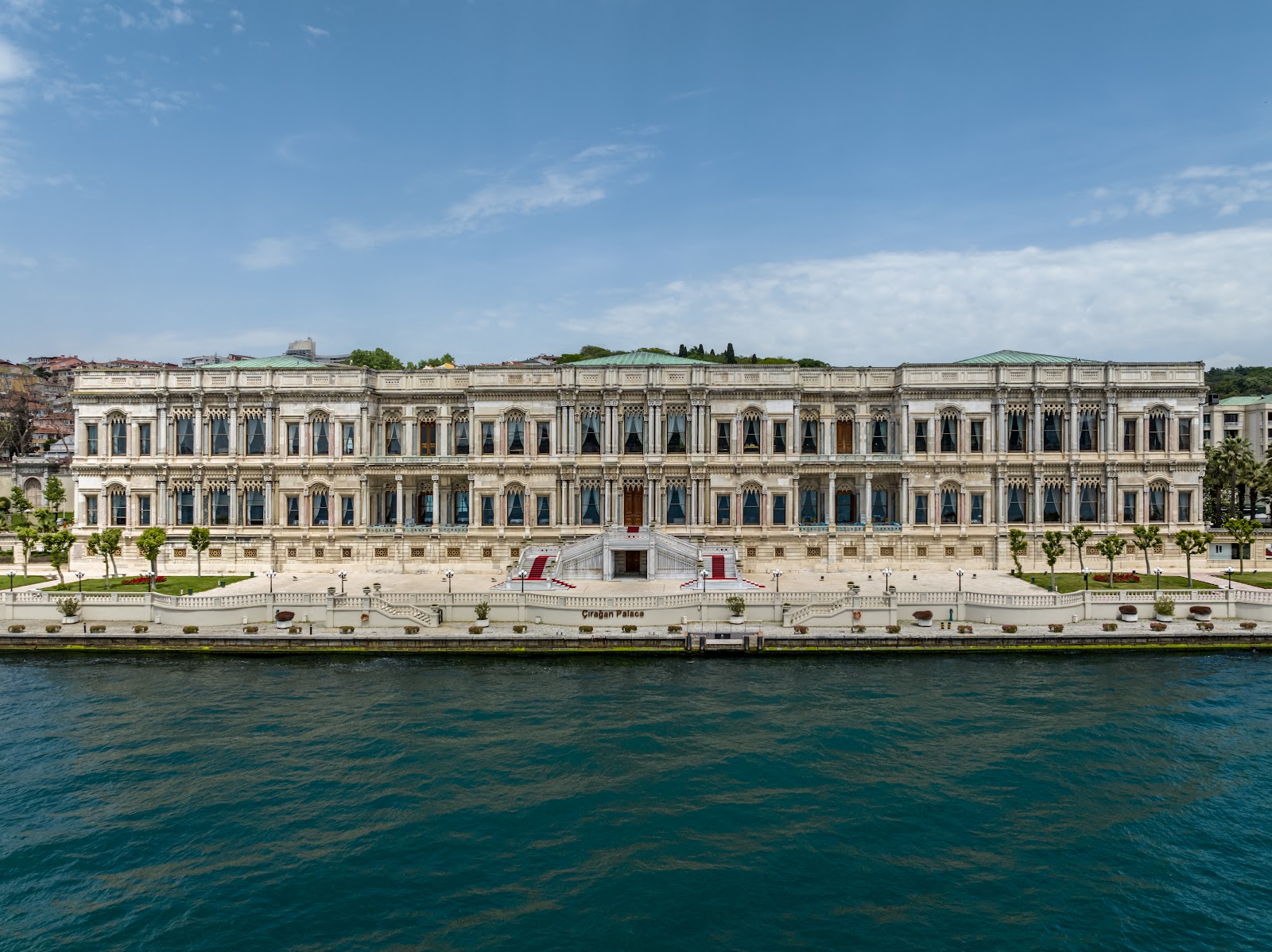 The symmetrical and grand facade of Ciragan Palace Kempinski seen from the Bosphorus on a bright, sunny day.