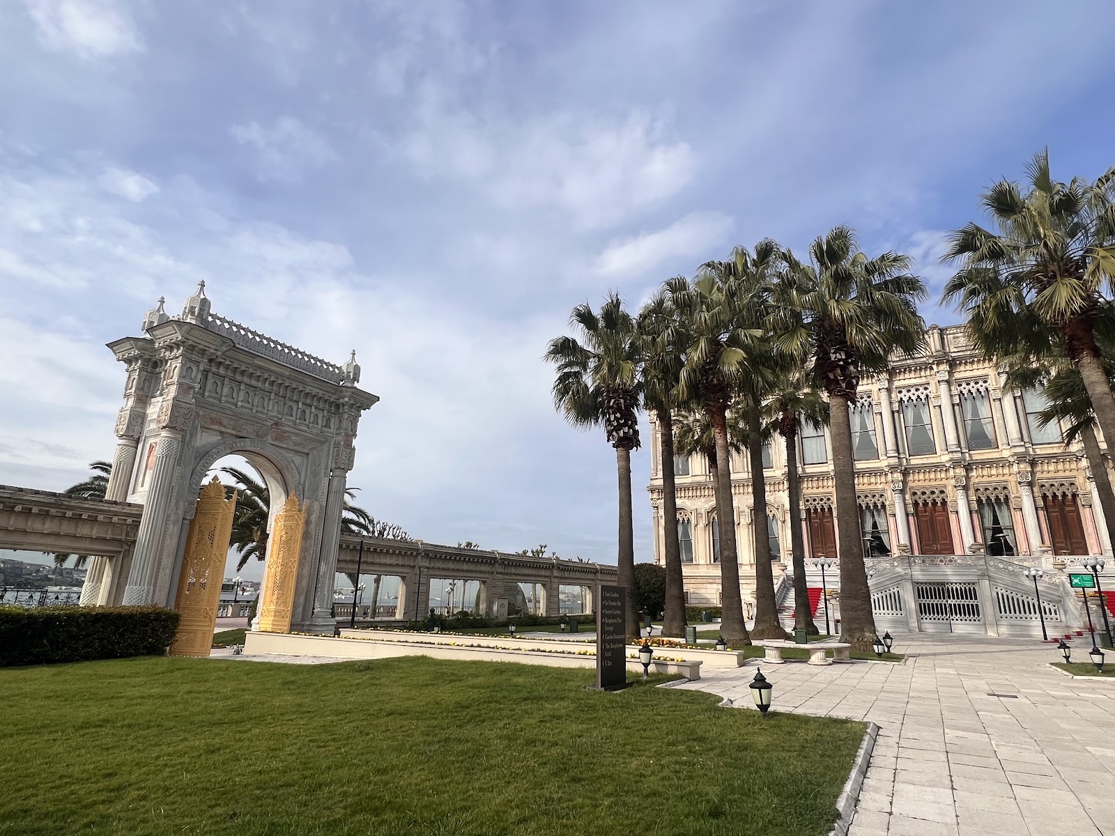 A ground-level view of Ciragan Palace Kempinski's elegant entrance, featuring a grand archway, tall palm trees, and manicured lawns.