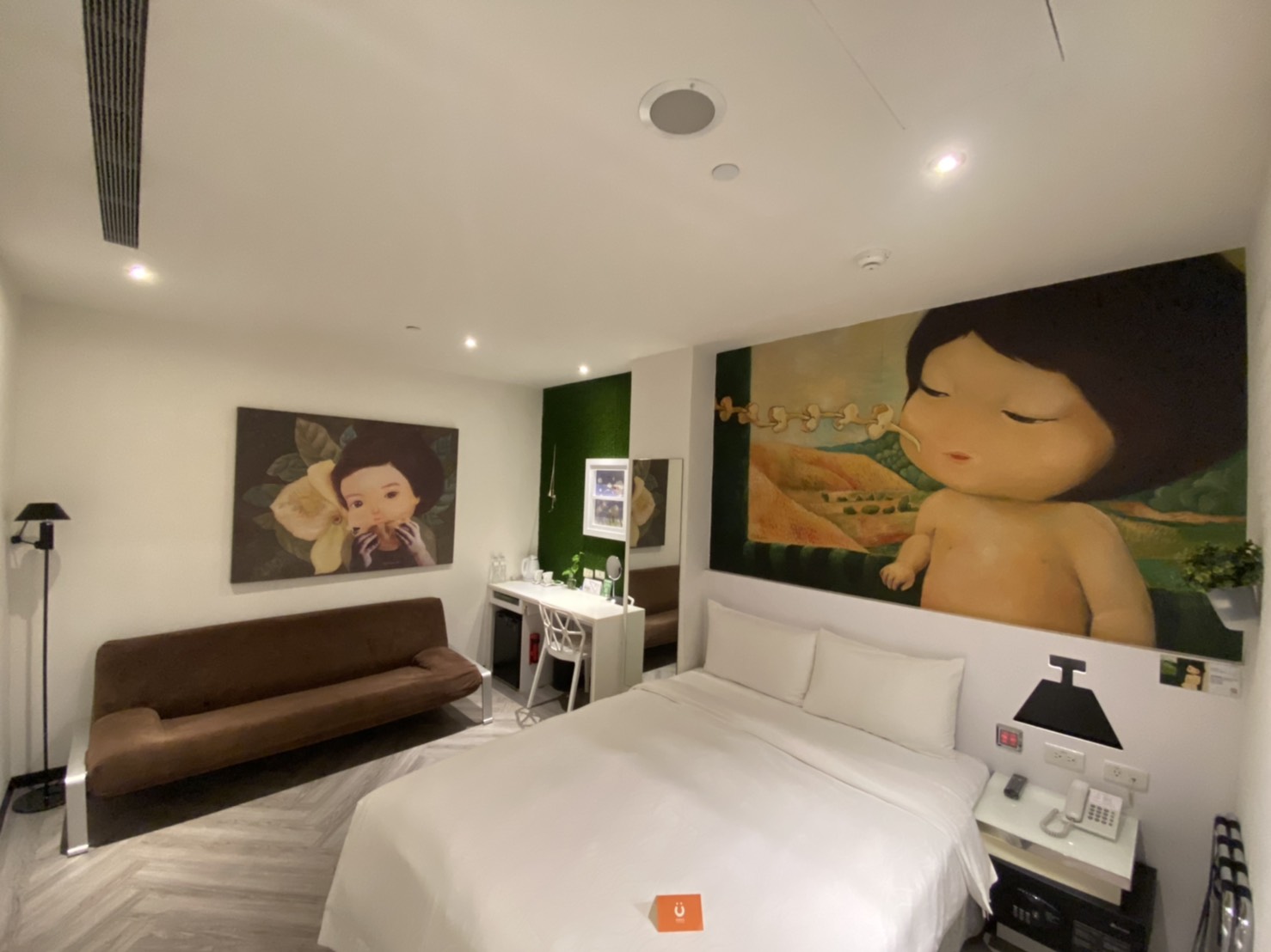 A cozy guest room with a queen-size bed, a brown sofa bed, a desk area, and large, whimsical wall art depicting a stylized figure.