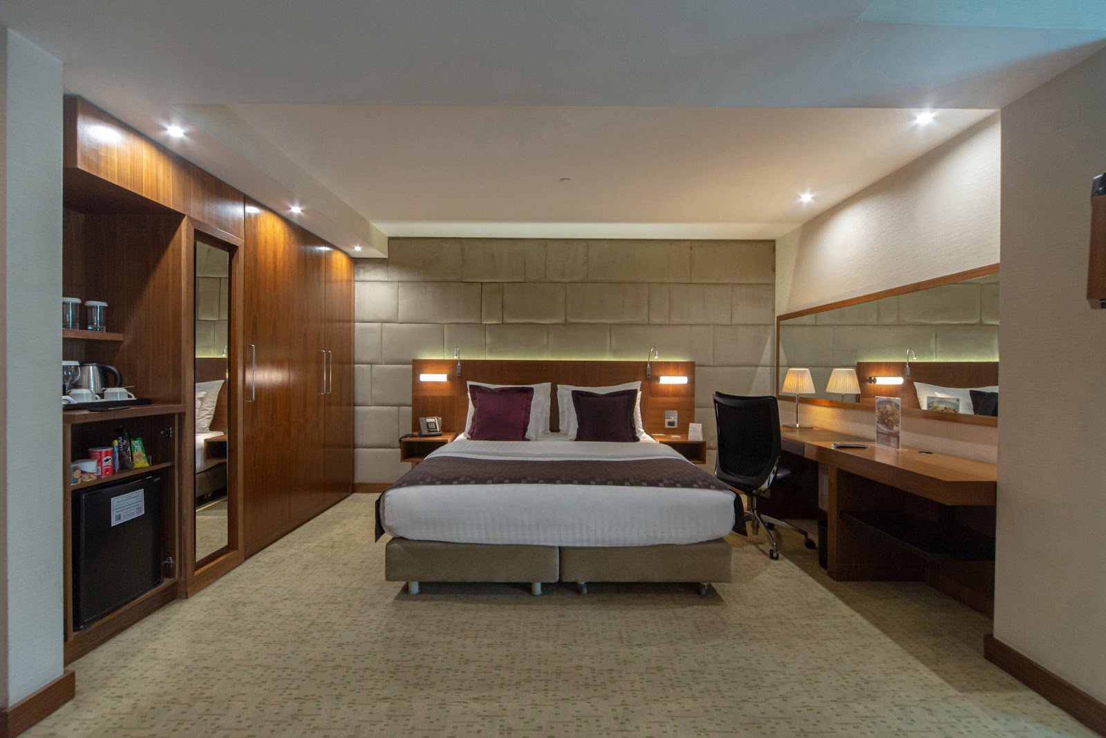 A modern hotel room with a queen-sized bed, a built-in wardrobe, a mini-bar, and a desk.