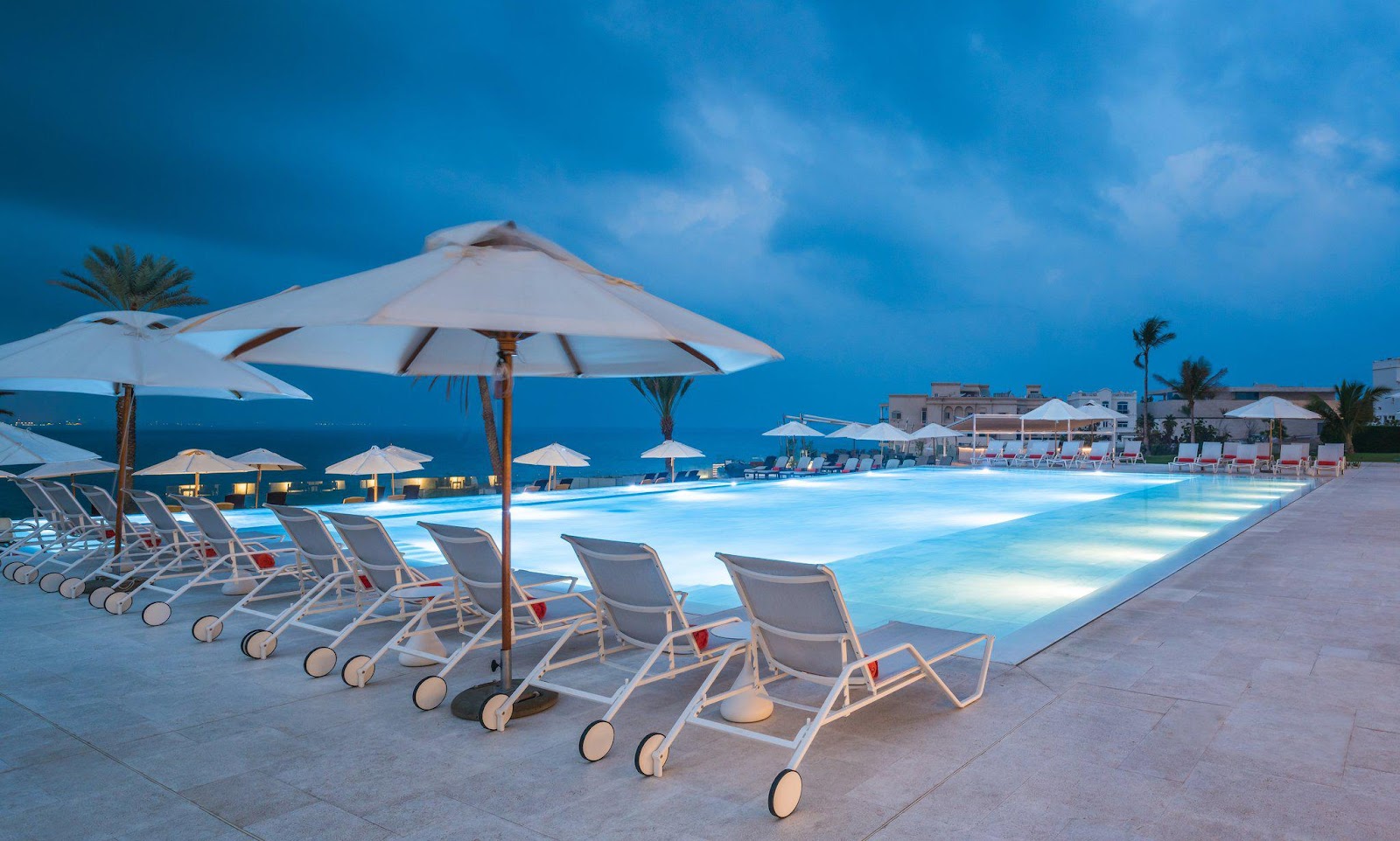 An illuminated outdoor swimming pool at night, surrounded by sun loungers and umbrellas, with the sea visible in the distance under a dramatic sky.