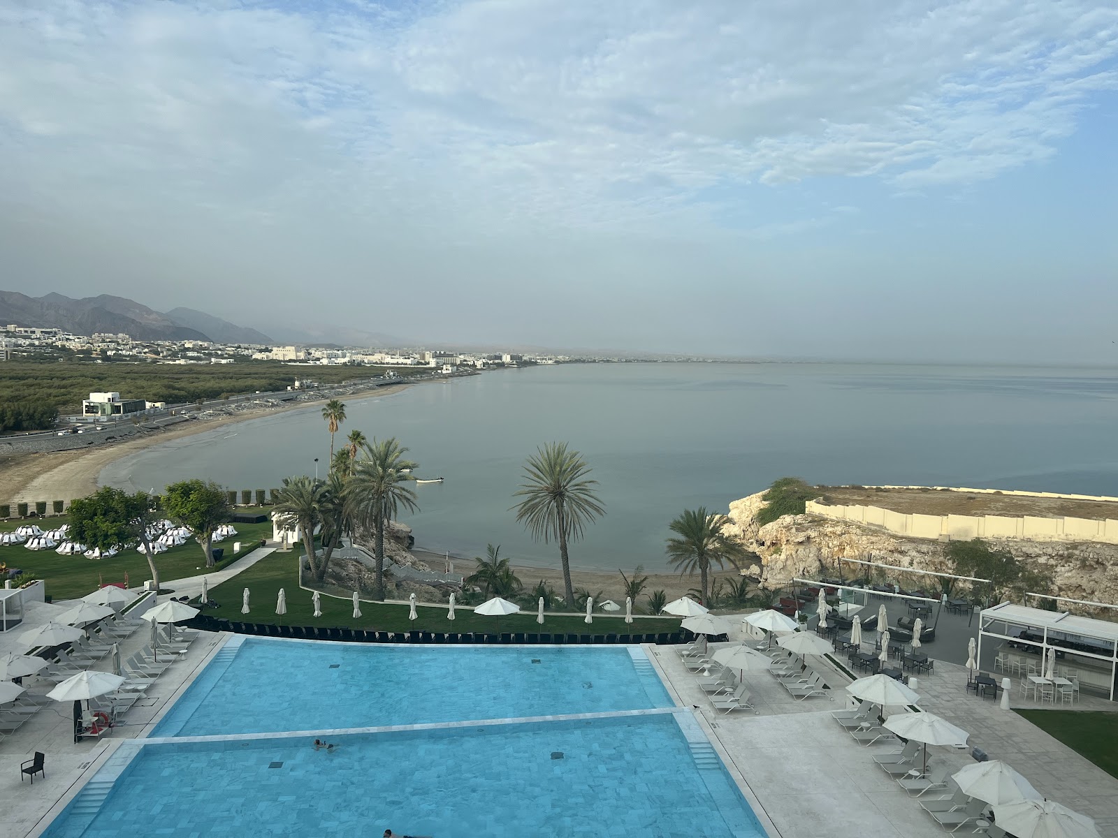 An expansive outdoor swimming pool surrounded by sun loungers and umbrellas, with a view of the beach, sea, and distant cityscape under a cloudy sky.