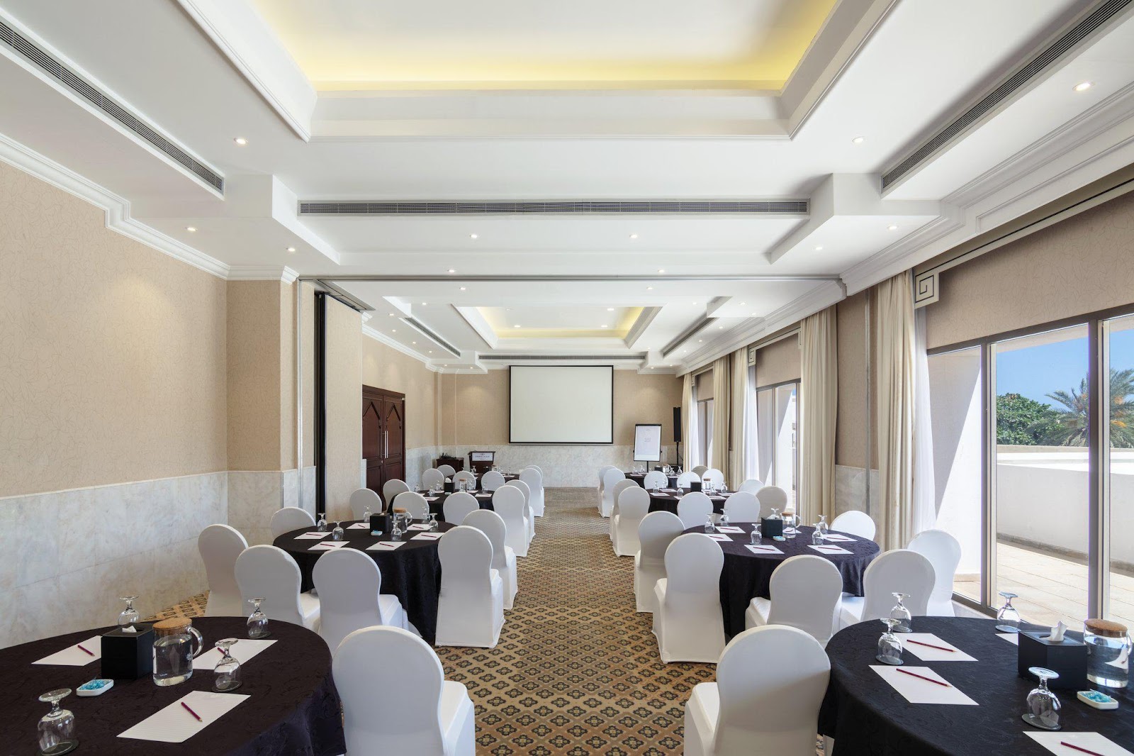 A bright conference or meeting room set up with round tables and chairs, a projector screen, and access to an outdoor balcony.