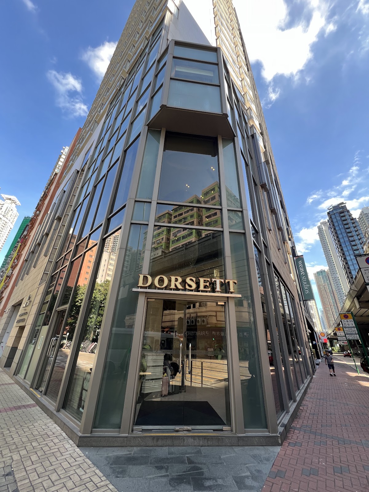 A low-angle shot of the Dorsett Mongkok hotel's modern glass facade, highlighting its contemporary architecture against a blue sky with clouds. A pedestrian is visible in the distance.