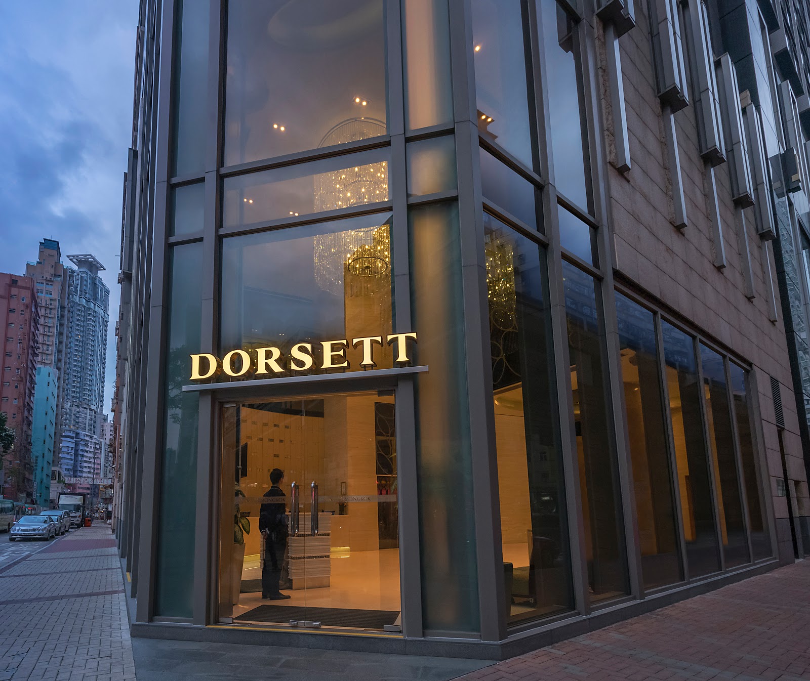 Another view of the Dorsett Mongkok hotel entrance from the street level, capturing the modern glass facade and the hotel sign. A person is seen standing inside the entrance.