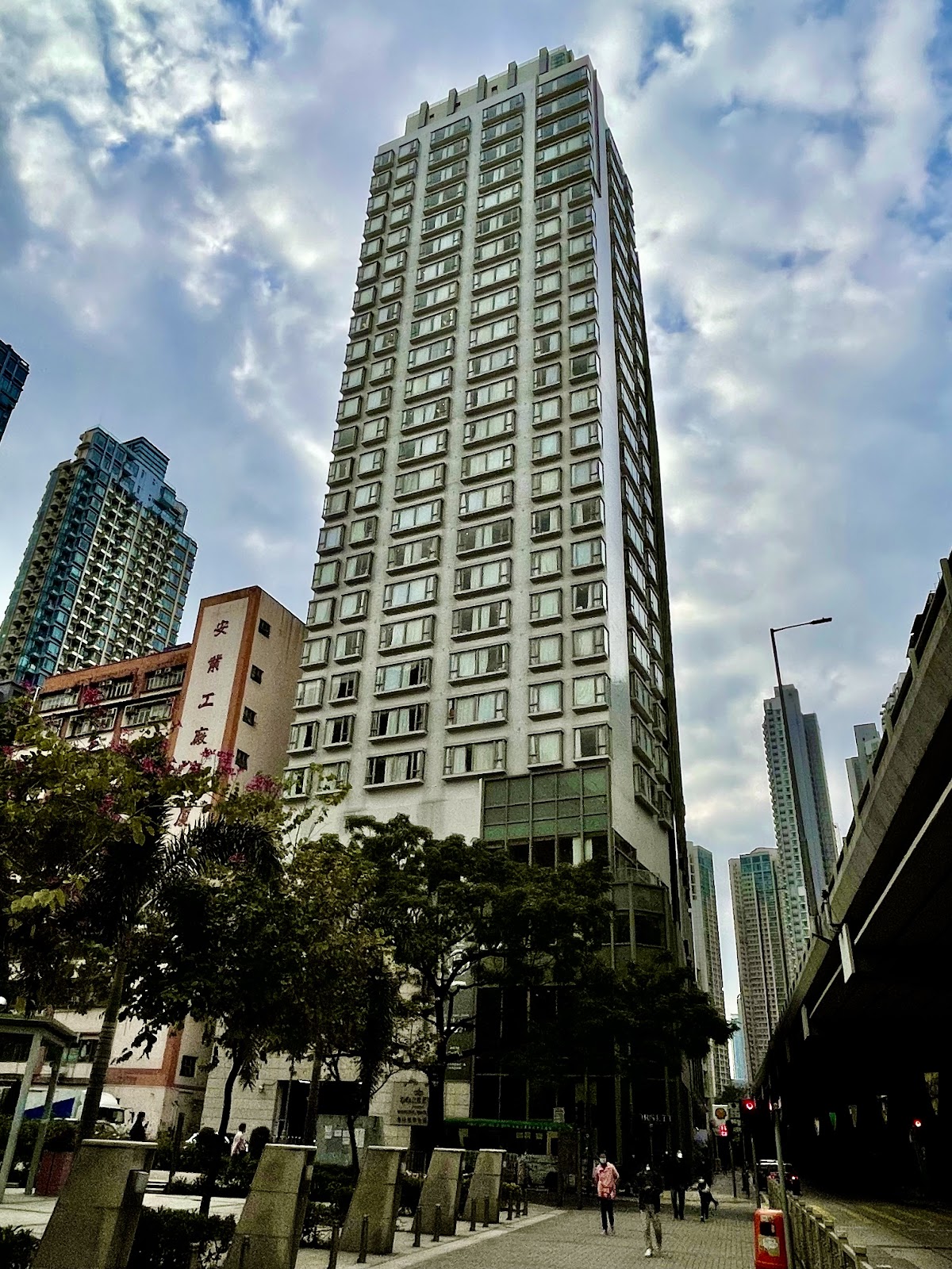 A full, tall shot of the Dorsett Mongkok hotel building, showcasing its slender, high-rise structure amidst the dense urban landscape of Hong Kong.