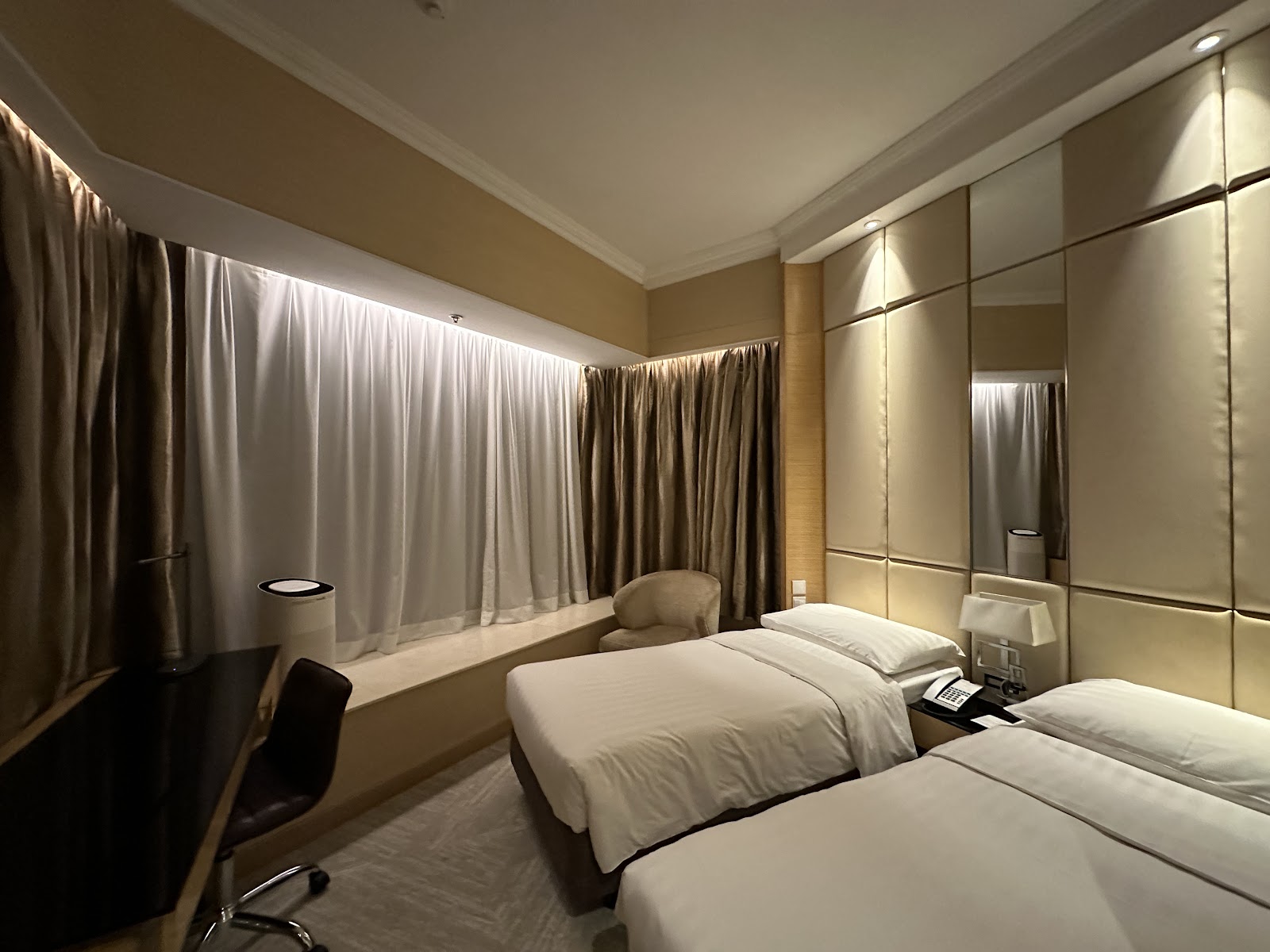A cozy hotel room featuring two single beds, a desk with a chair, and full-length curtains covering a large window. The room has warm lighting and modern decor.