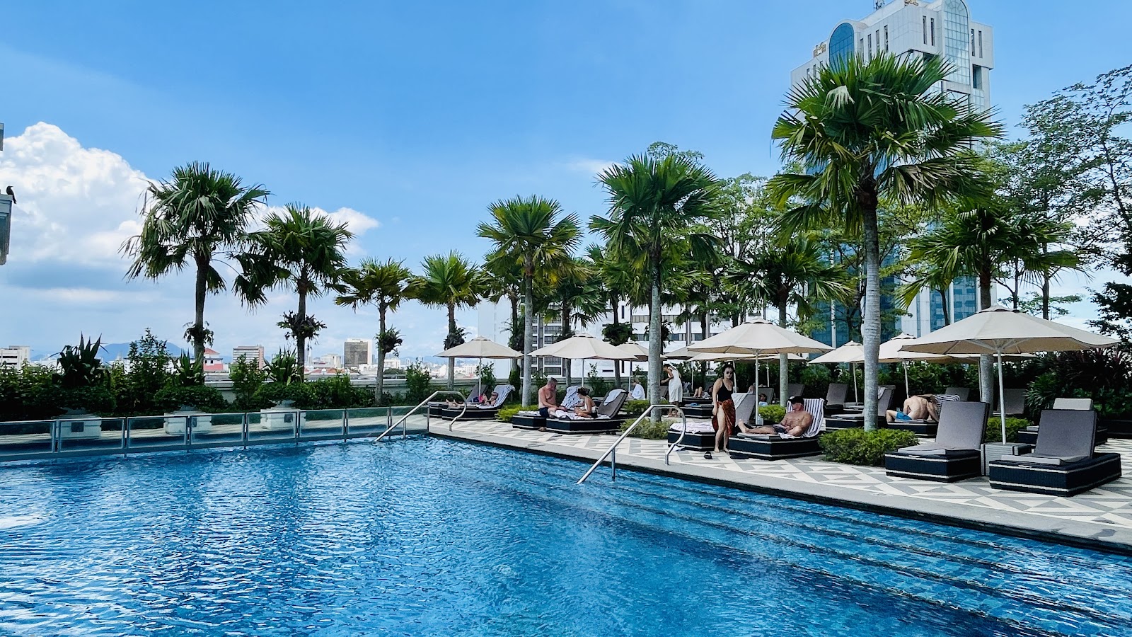 A vibrant outdoor shared infinity pool area with numerous sun loungers, lush palm trees, and a distant city skyline under a clear blue sky. Several guests are visible in swimwear.