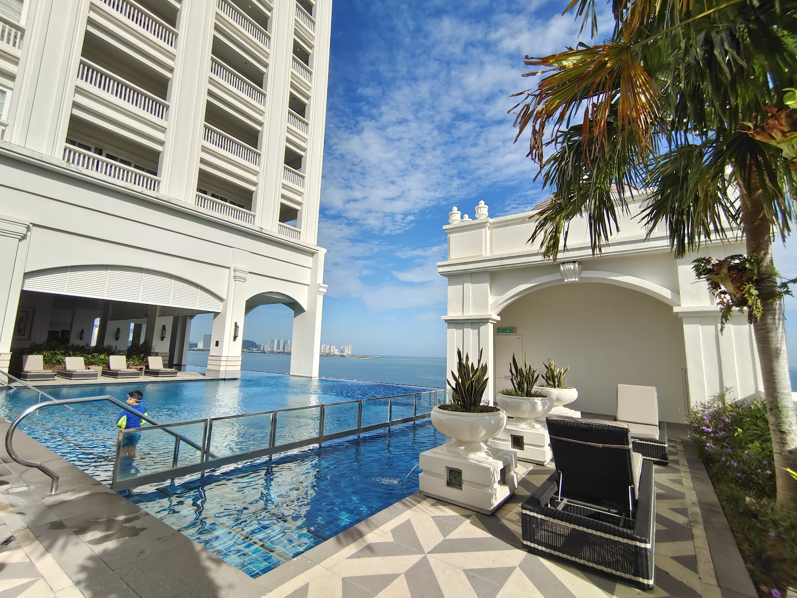 Another view of an outdoor infinity pool, possibly a different section or a second pool, situated beside a modern hotel building. It overlooks the sea and features a child swimming.