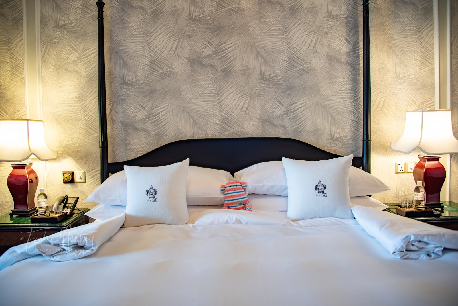 A luxurious bedroom showcasing a four-poster king-size bed with elegant linens and throw pillows, complemented by stylish side lamps and tropical patterned wallpaper.