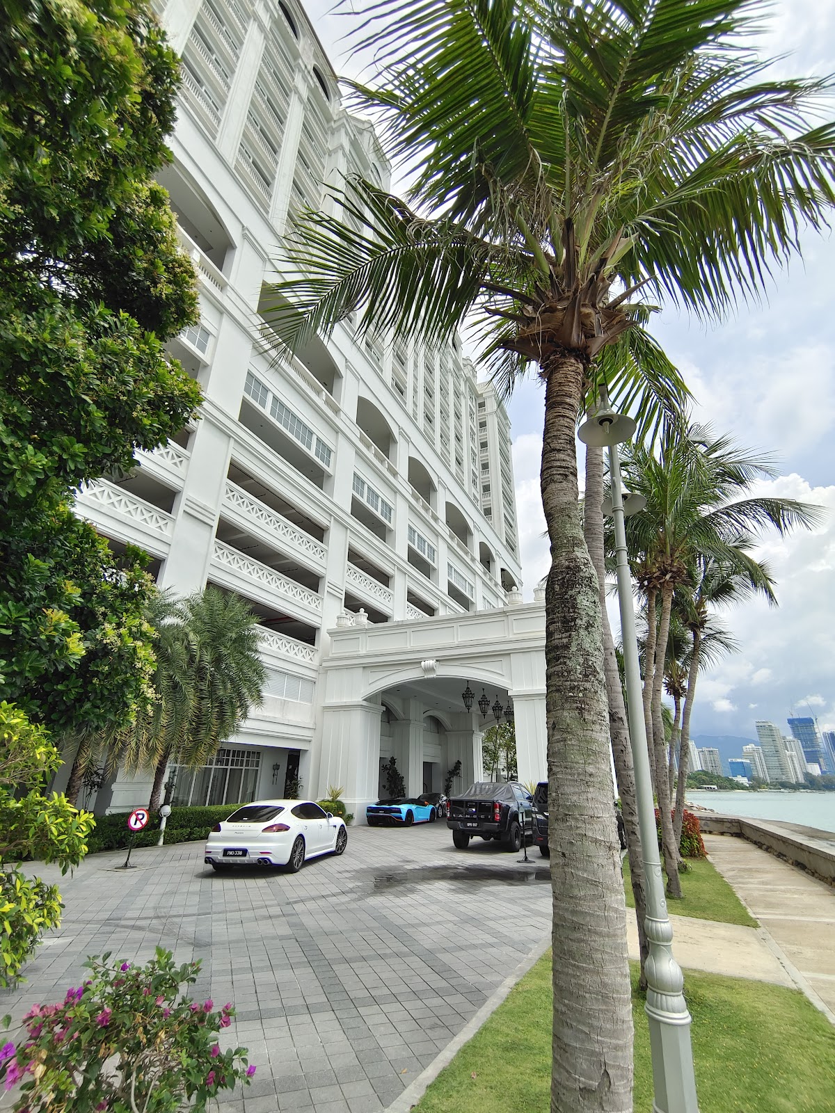 The grand entrance of the modern wing of the Eastern & Oriental Hotel, featuring a stylish porte-cochère, luxury cars, tall palm trees, and classic architectural details.