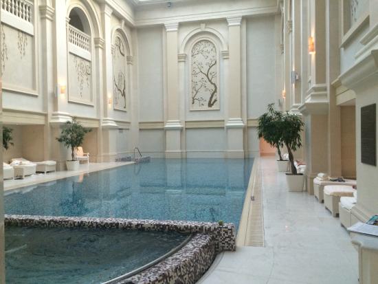 An opulent indoor swimming pool area with classical architectural elements, high ceilings, and comfortable seating alcoves, currently empty and well-lit.