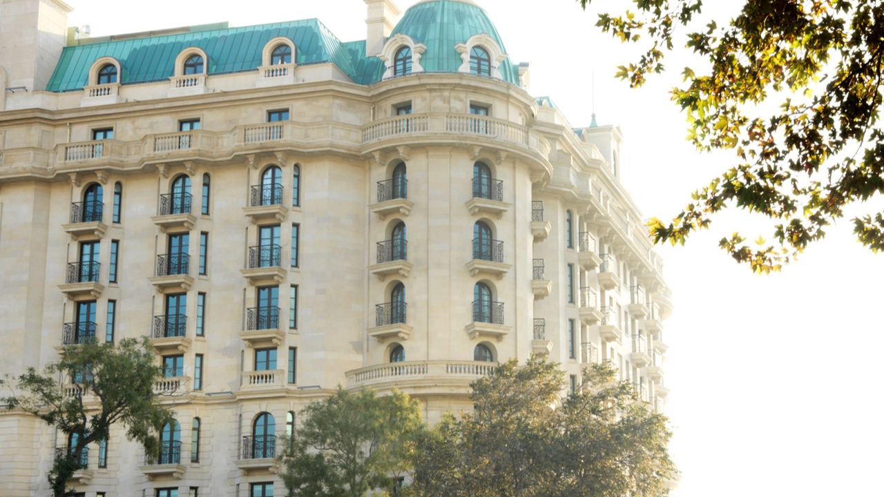 A close-up view of the Four Seasons Hotel Baku's elegant facade with its distinctive arched windows and light-colored stone, partially framed by green foliage under bright sunlight.