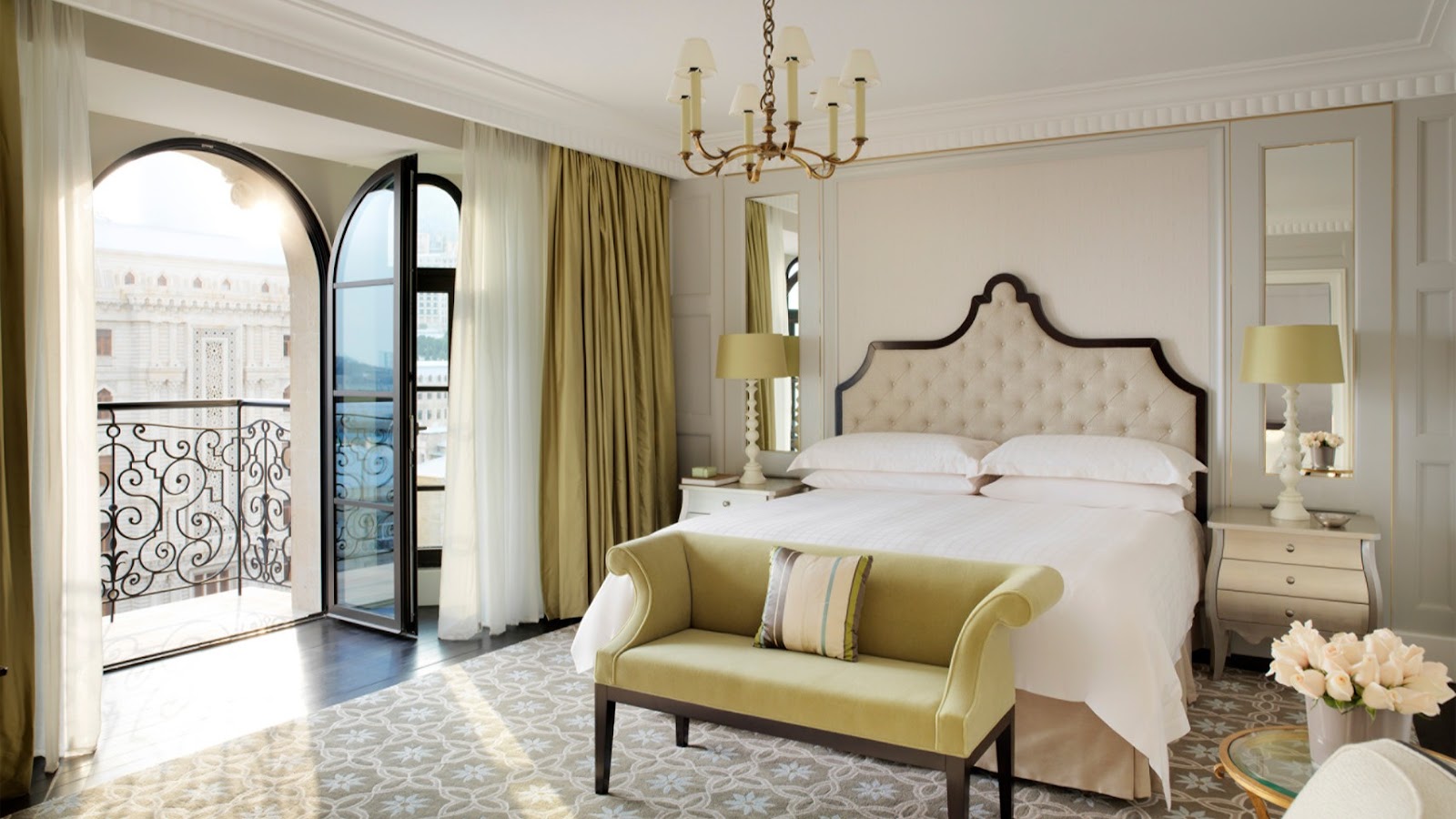 A luxurious hotel room featuring a king-size bed, elegant headboard, a cozy seating bench, and a balcony with an ornate railing offering a view of the city's classic architecture.