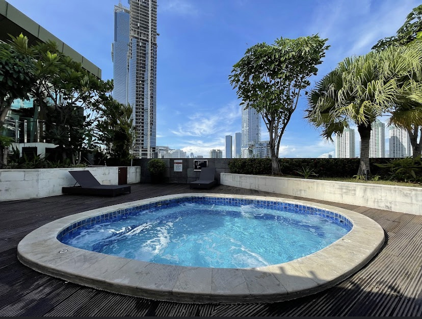 An outdoor rooftop jacuzzi or small plunge pool with bubbling water, surrounded by wooden decking and high-rise city buildings in the background.