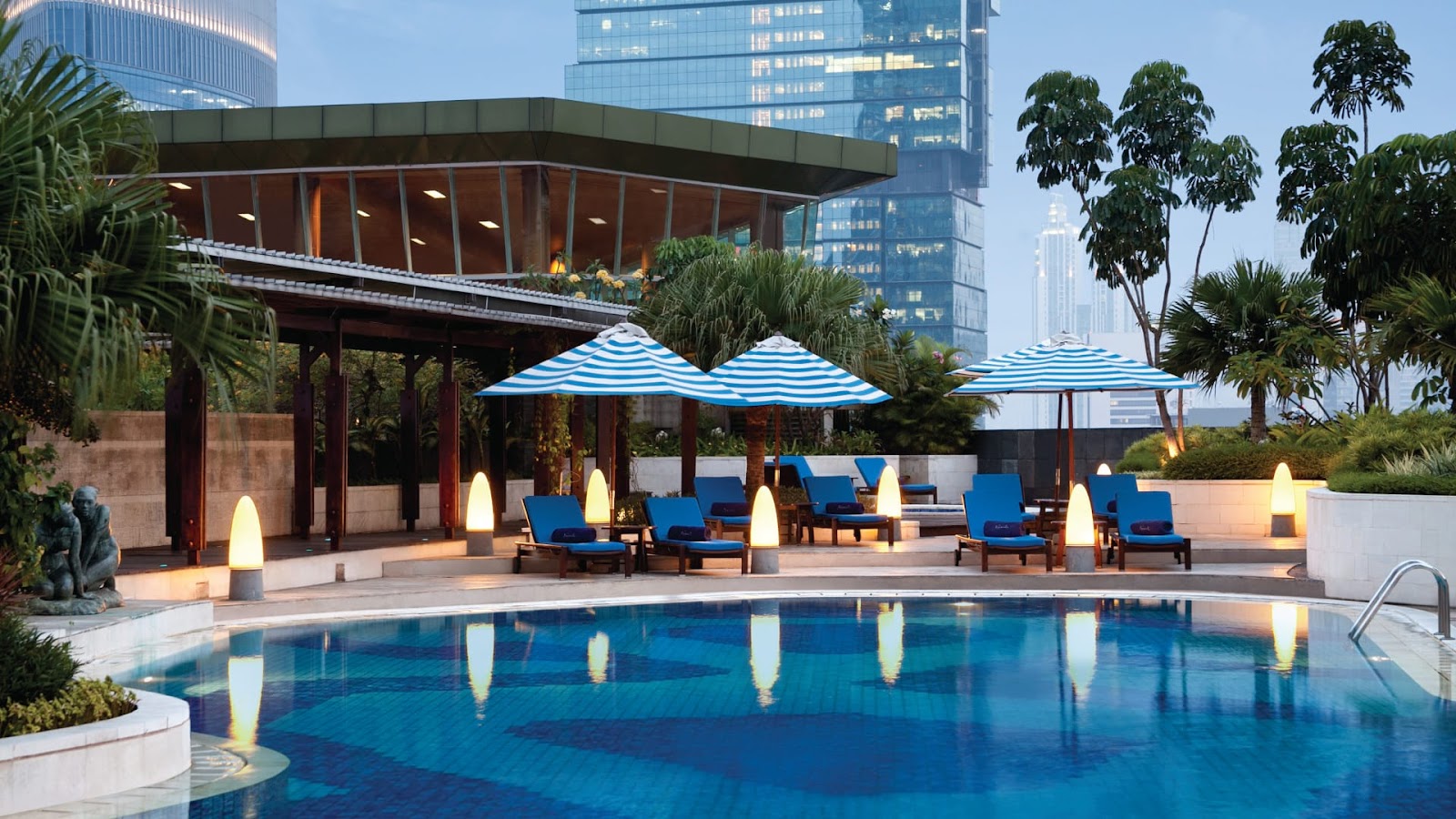 A large, beautifully lit outdoor swimming pool on a landscaped terrace, surrounded by sun loungers with blue umbrellas and tropical greenery, seen at dusk.