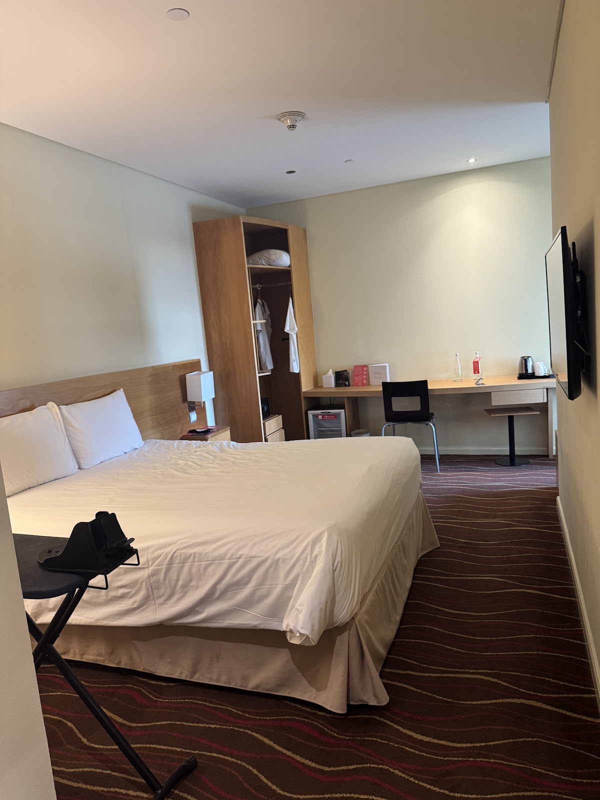 A comfortable hotel room featuring a queen-sized bed, a desk area with a chair, a wall-mounted TV, and an open wardrobe.