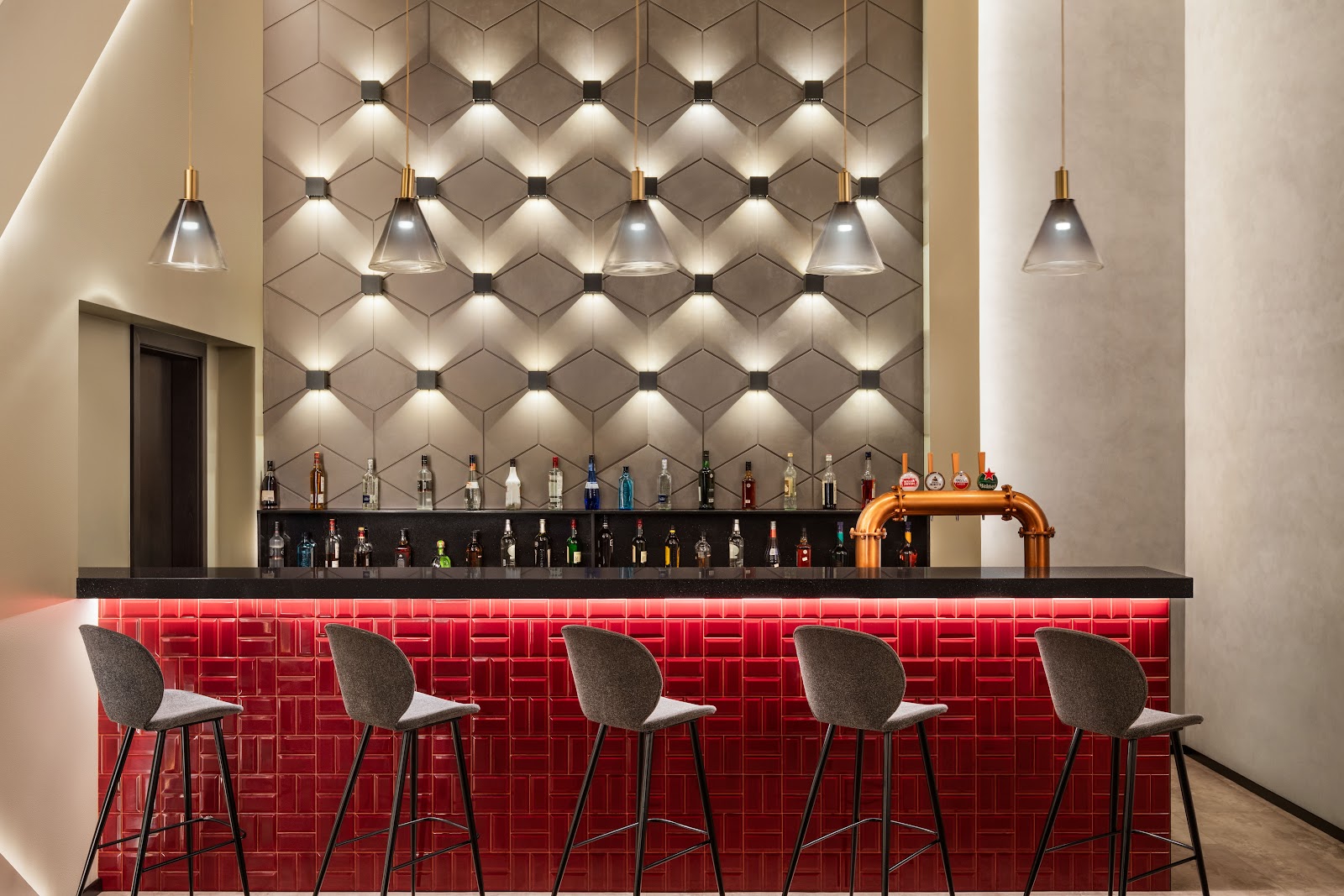 A modern bar area with a long red tiled counter, high stools, a backlit display of bottles, and an industrial-chic lighting design.