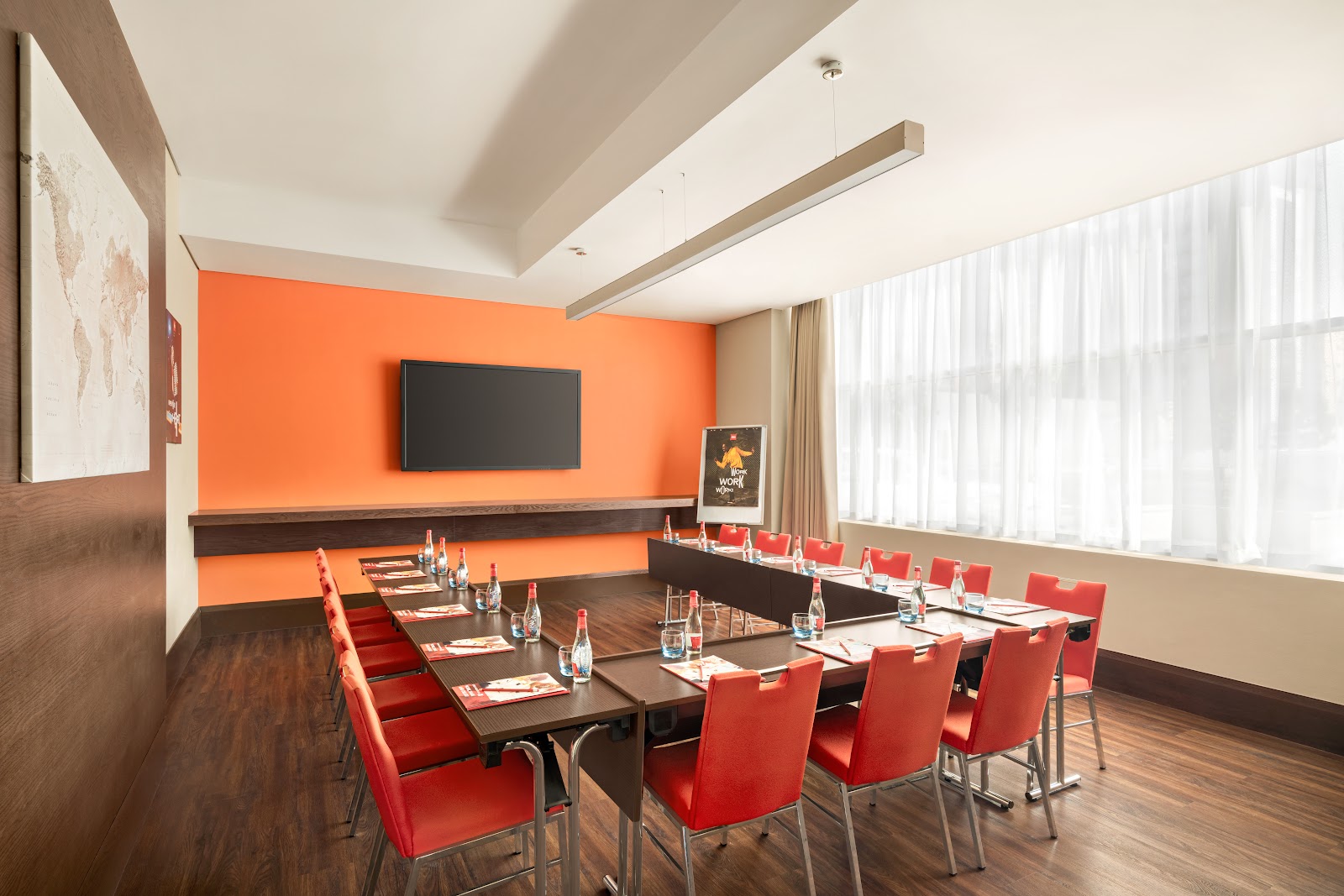 A spacious meeting room set up in a U-shape configuration with red chairs, a large wall-mounted screen, and bright orange accent walls.