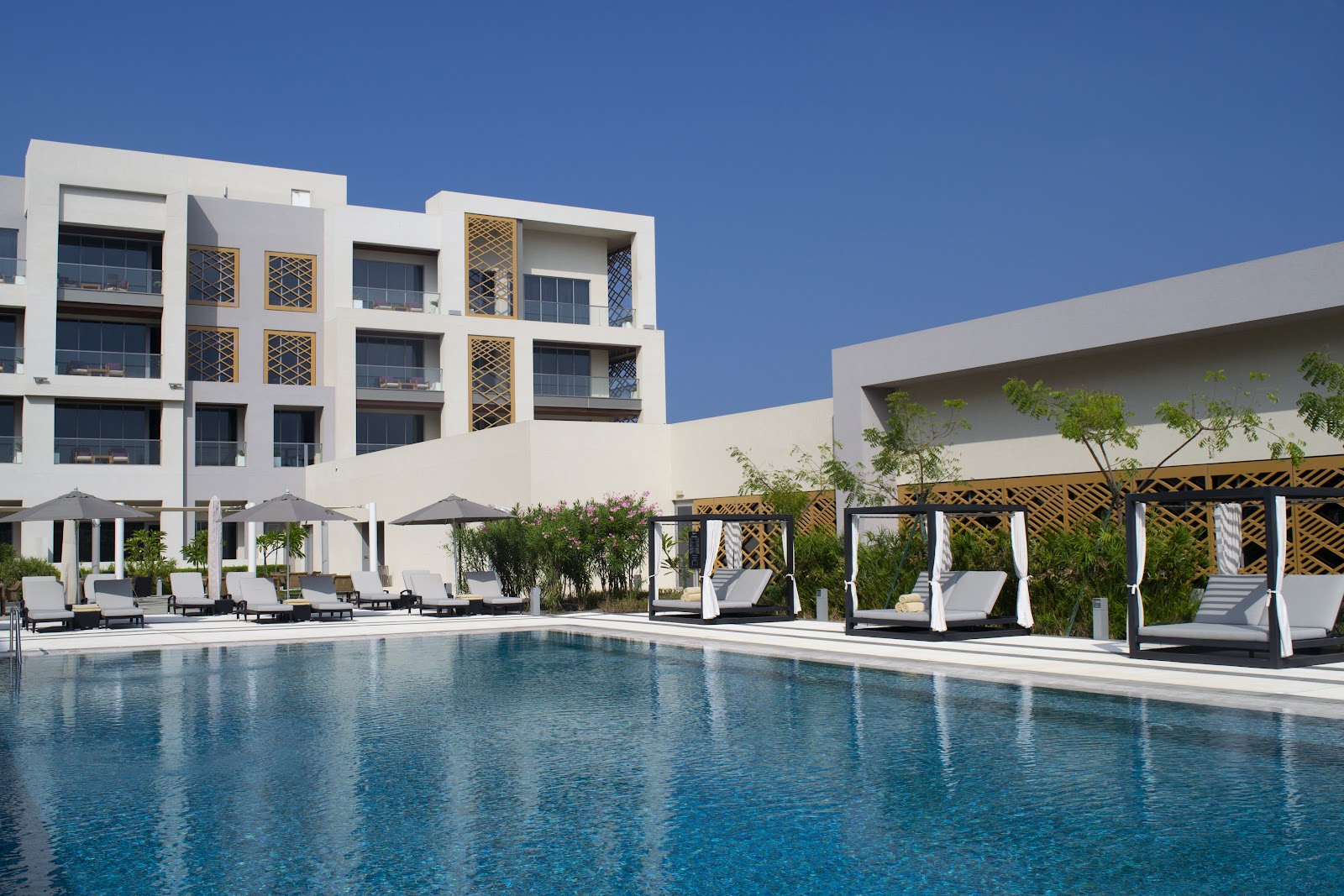 A large outdoor shared swimming pool with sun loungers and cabanas, facing a modern hotel building.