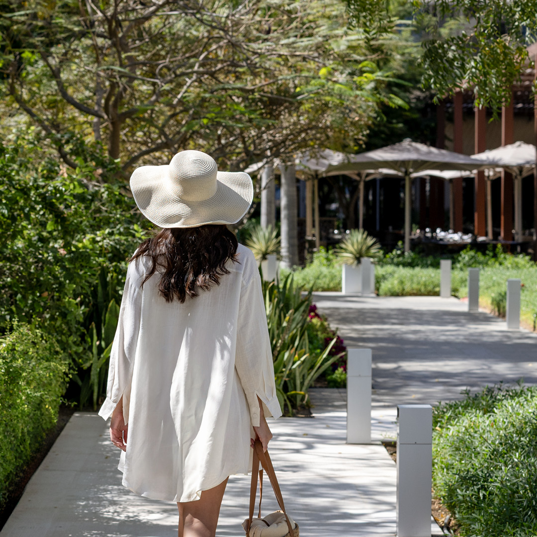 A guest walking along a lush pathway through resort gardens, with outdoor dining areas visible in the background.