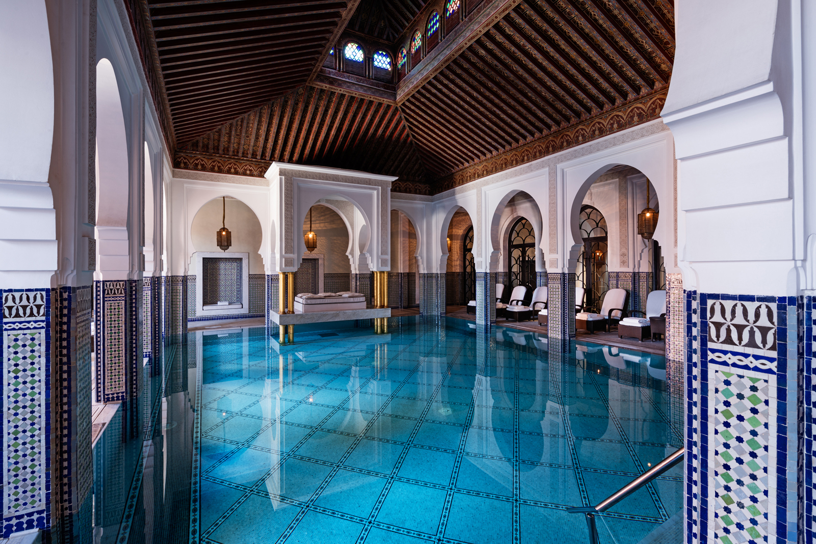A magnificent indoor swimming pool with traditional Moroccan arches, intricate blue and white tiling, a high wooden ceiling, and ambient lighting.