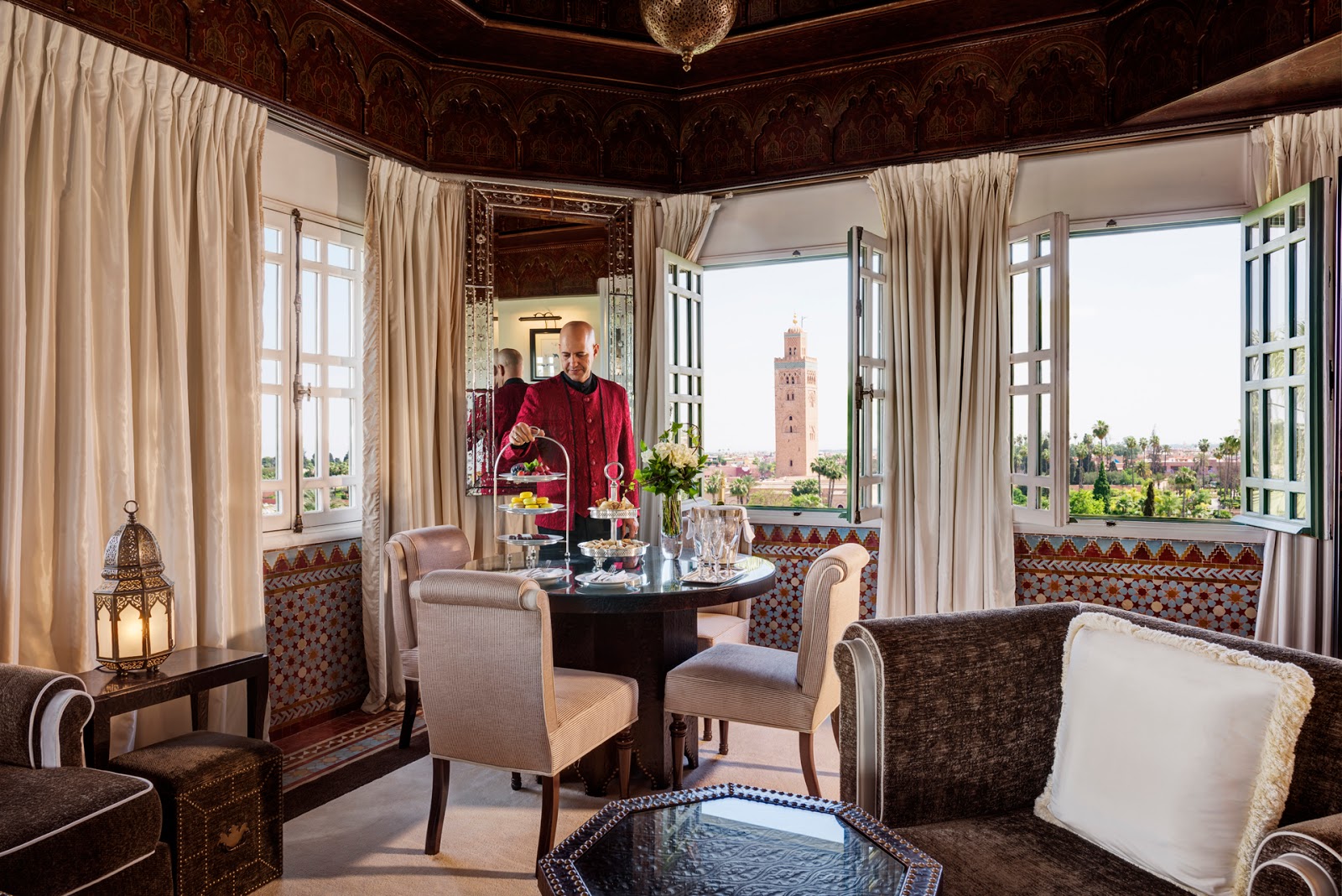 A luxurious suite with traditional Moroccan decor, a dining table set for tea, and an open window offering a direct view of the Koutoubia Mosque.