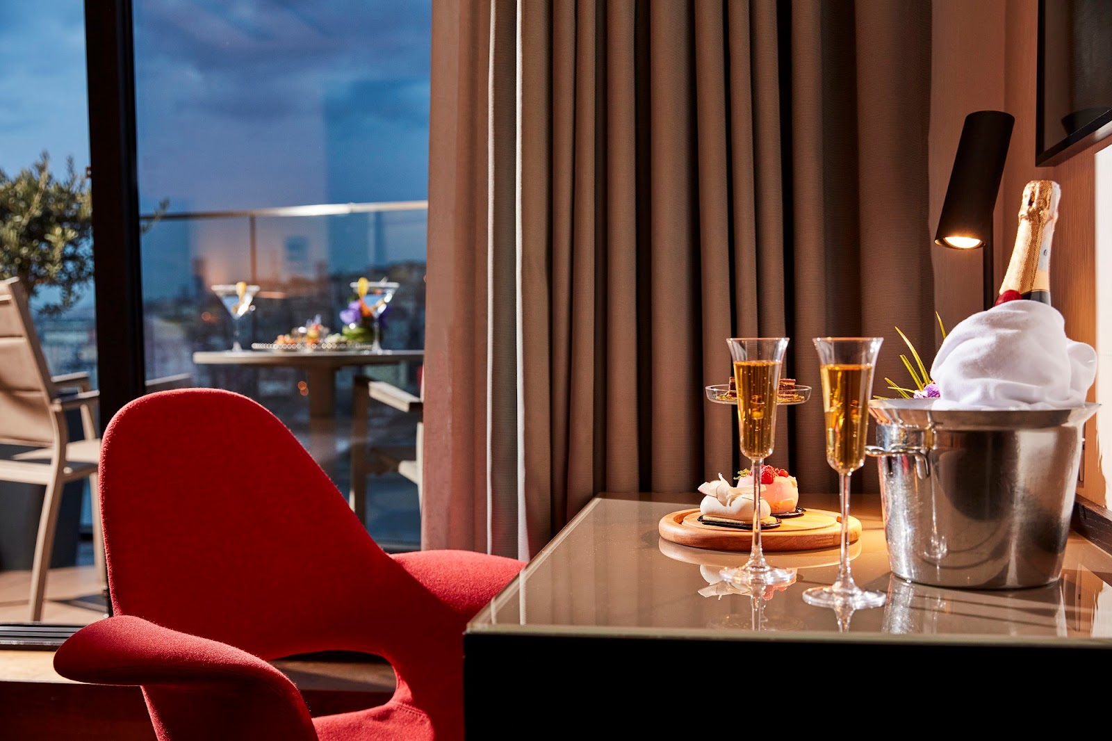 An intimate corner of a hotel room with a desk, a red chair, and a balcony view with drinks and snacks, suggesting a welcome amenity or in-room dining.