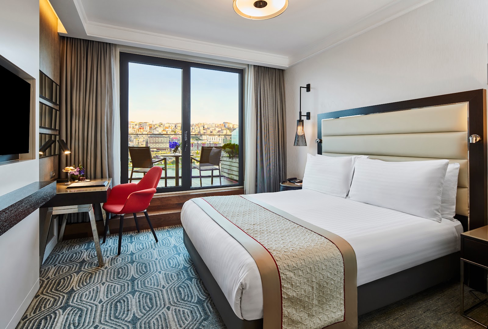 A stylish hotel room featuring a king-size bed, a comfortable work desk with a red chair, and a private balcony overlooking the city.
