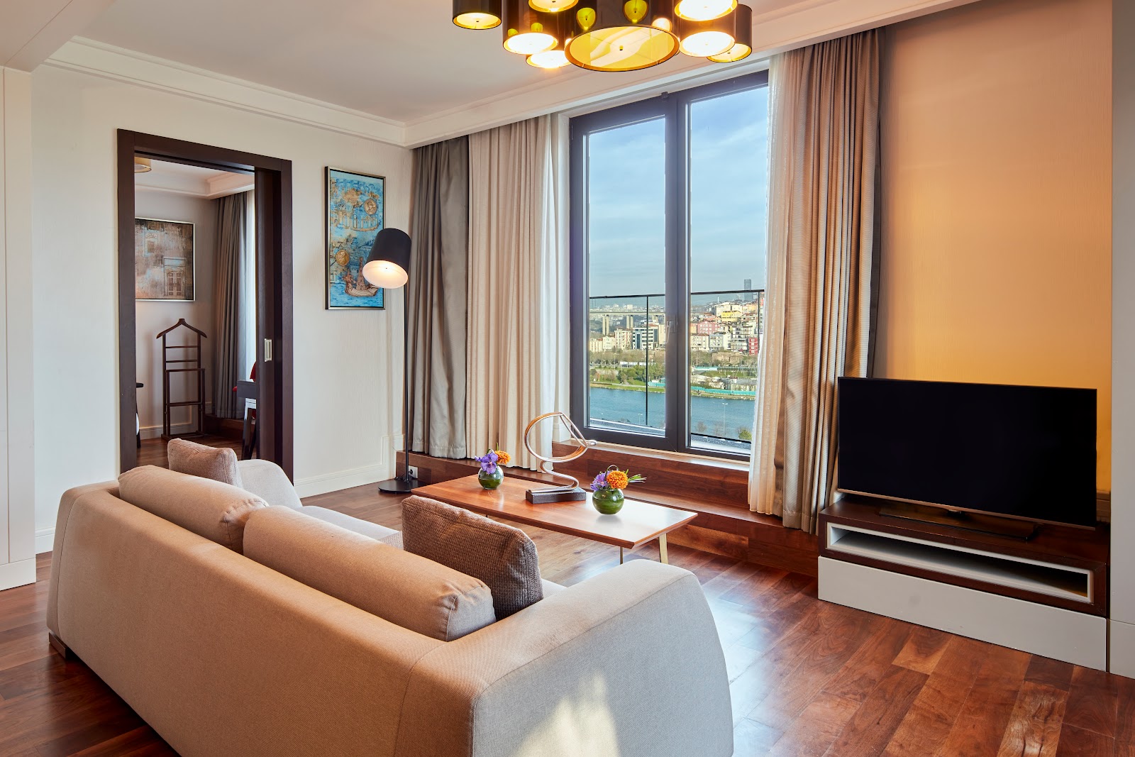 A spacious and elegantly furnished living area of a hotel suite, featuring a comfortable sofa, a TV, and large windows with city views.