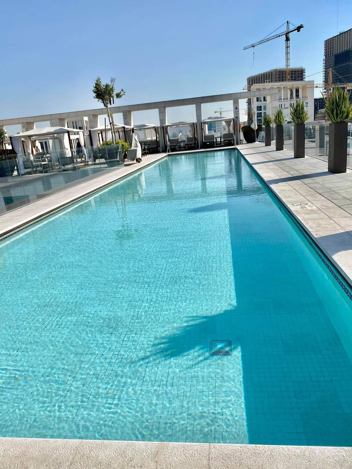 An empty outdoor rooftop swimming pool with sun loungers and cabanas, offering city views.
