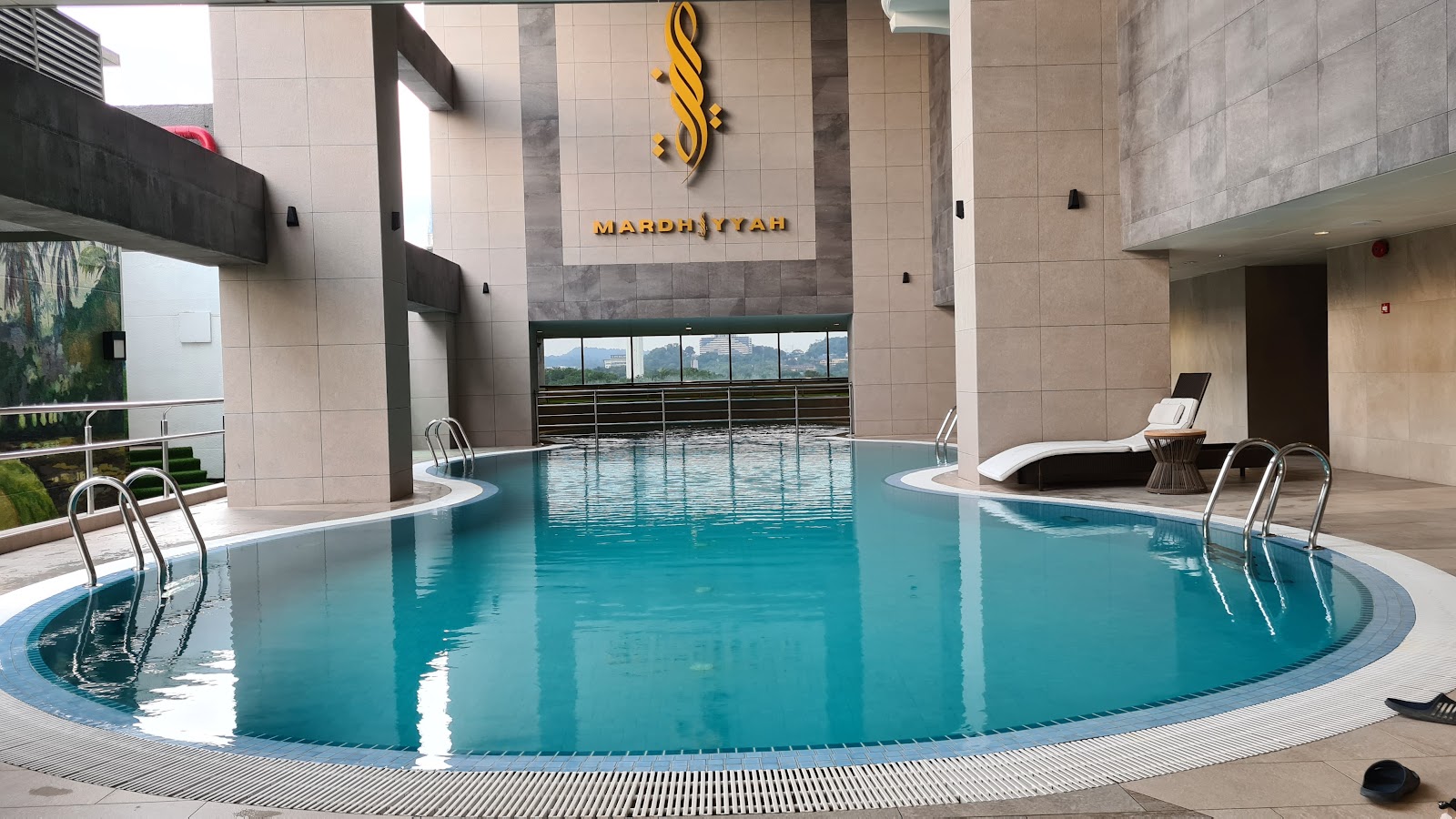 An outdoor swimming pool located on an upper floor, featuring lounge chairs and framed by hotel architecture, with a distant view of the city/hills.
