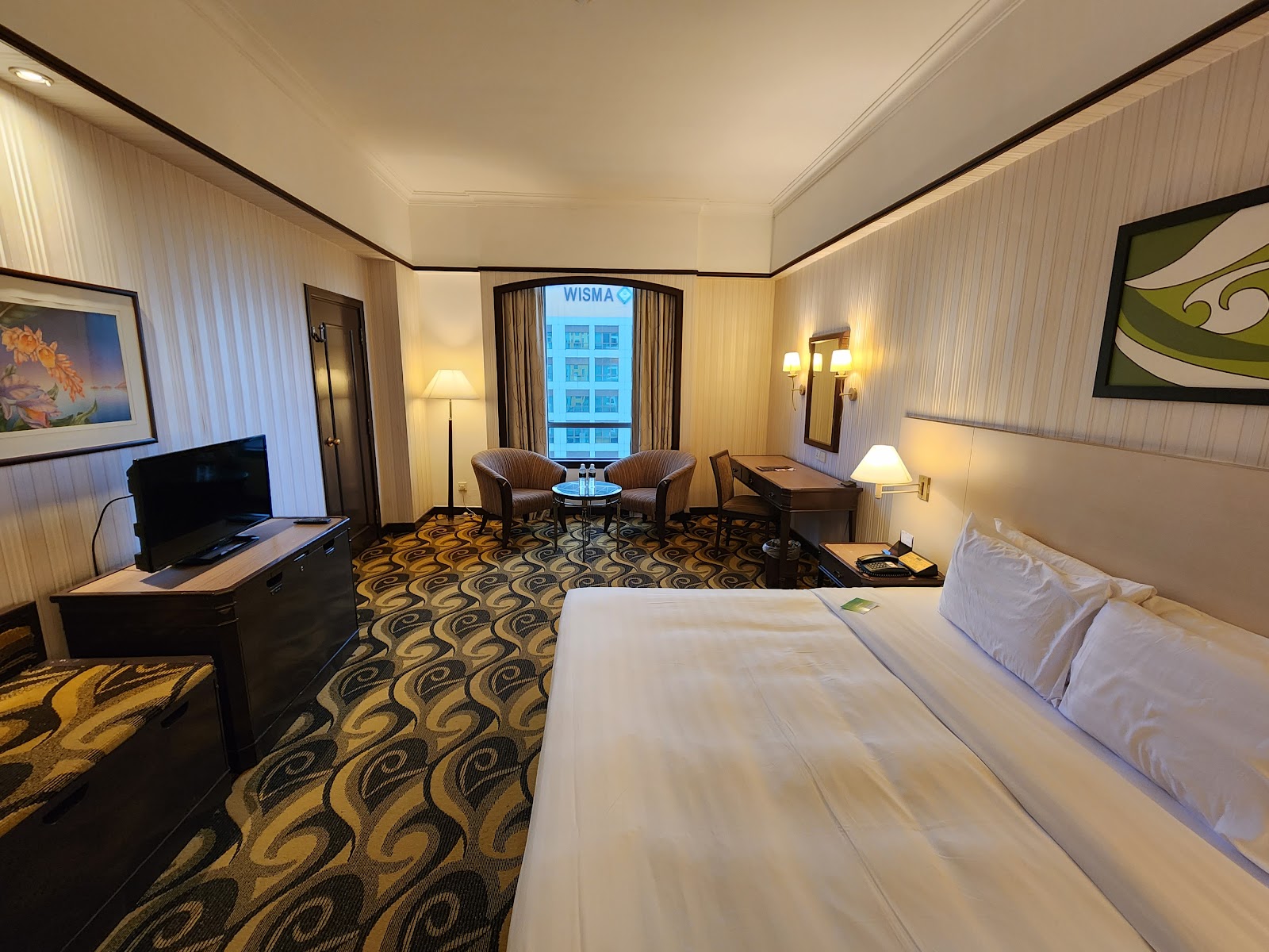 A well-appointed hotel room featuring a king-sized bed, a comfortable seating area by the window, a work desk, and patterned carpet.