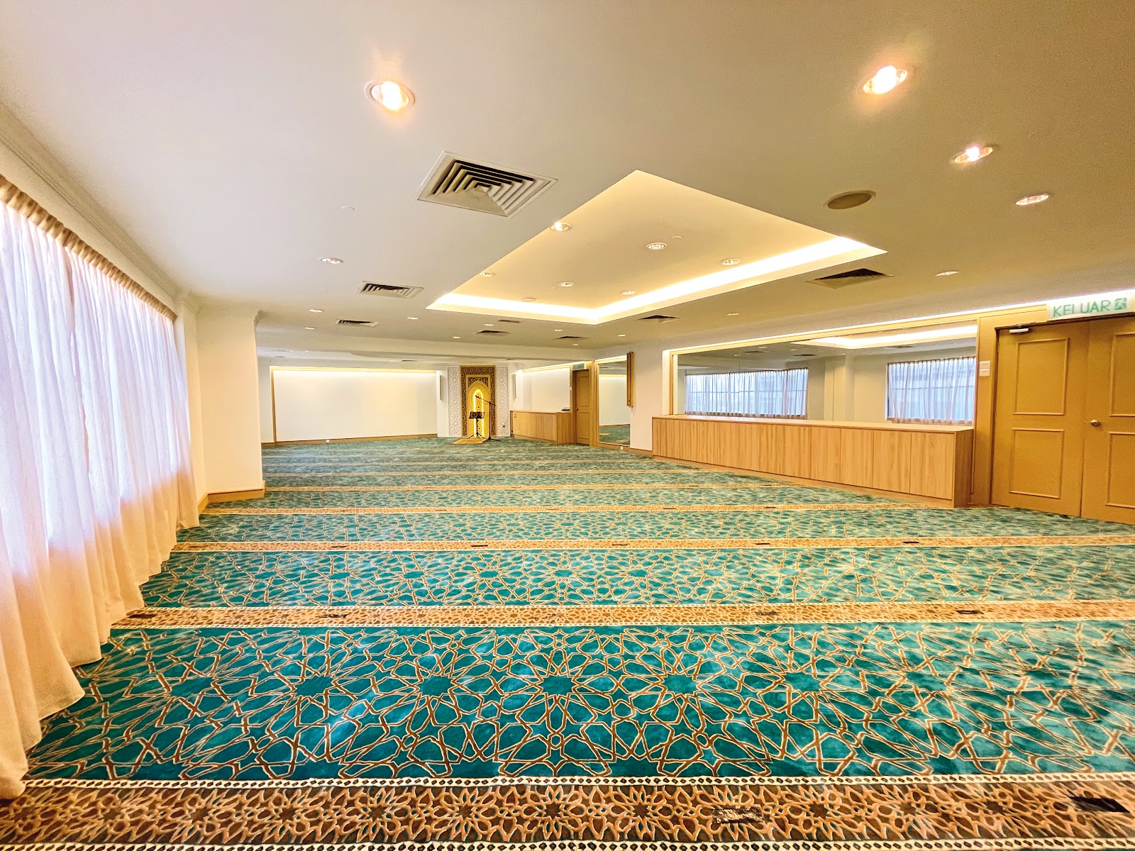 A spacious and elegantly carpeted prayer room (Musolla) with Islamic geometric patterns, a mihrab, and natural light, designed for guests' spiritual needs.