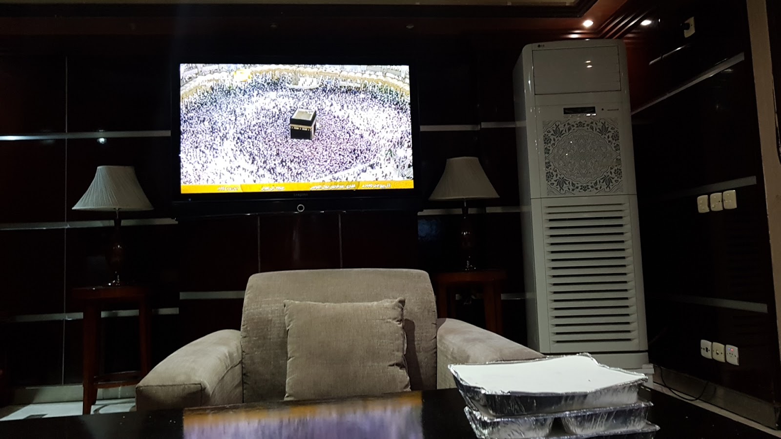 A lounge area with comfortable seating and a large TV displaying live coverage of the Kaaba, offering a tranquil space.