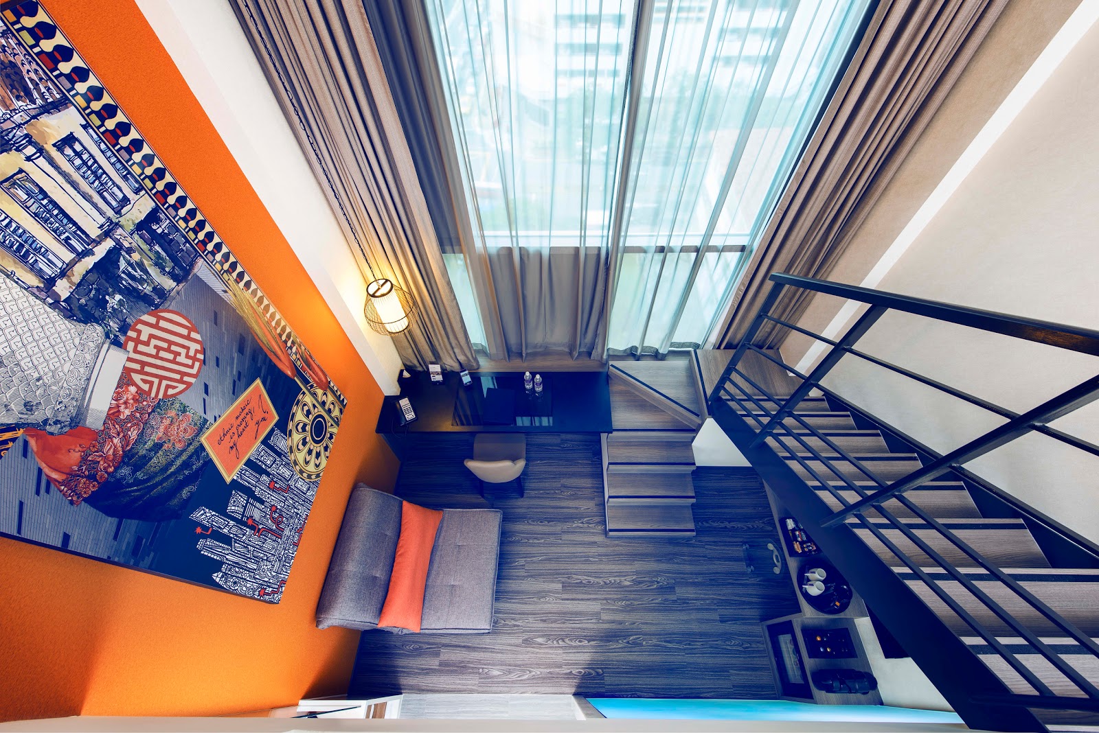 A unique loft-style hotel room viewed from above, showing a downstairs living area with a sofa and desk, and a staircase leading to an upper level.