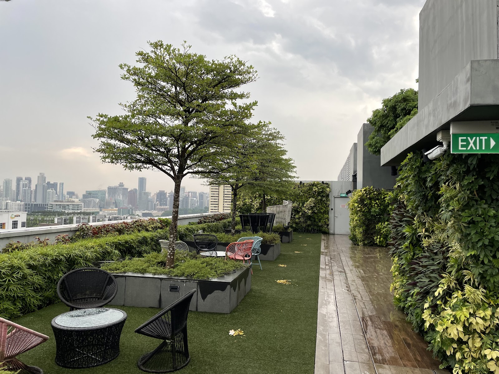 A vibrant rooftop garden lounge with seating areas, lush greenery, and a panoramic city skyline view under a cloudy sky.