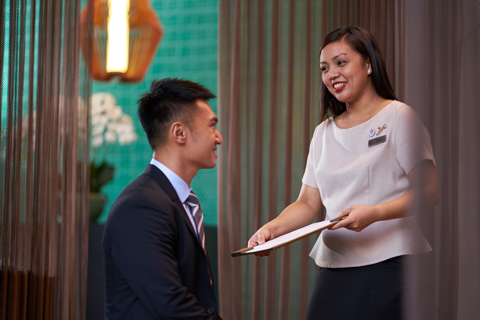 A hotel staff member (woman) professionally interacting with a guest (man) at a reception or concierge area.