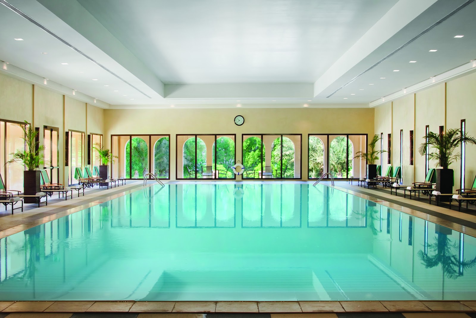 A large, bright indoor swimming pool within the hotel, featuring floor-to-ceiling arched windows that offer views of lush outdoor greenery. Lounge chairs are available around the pool area.