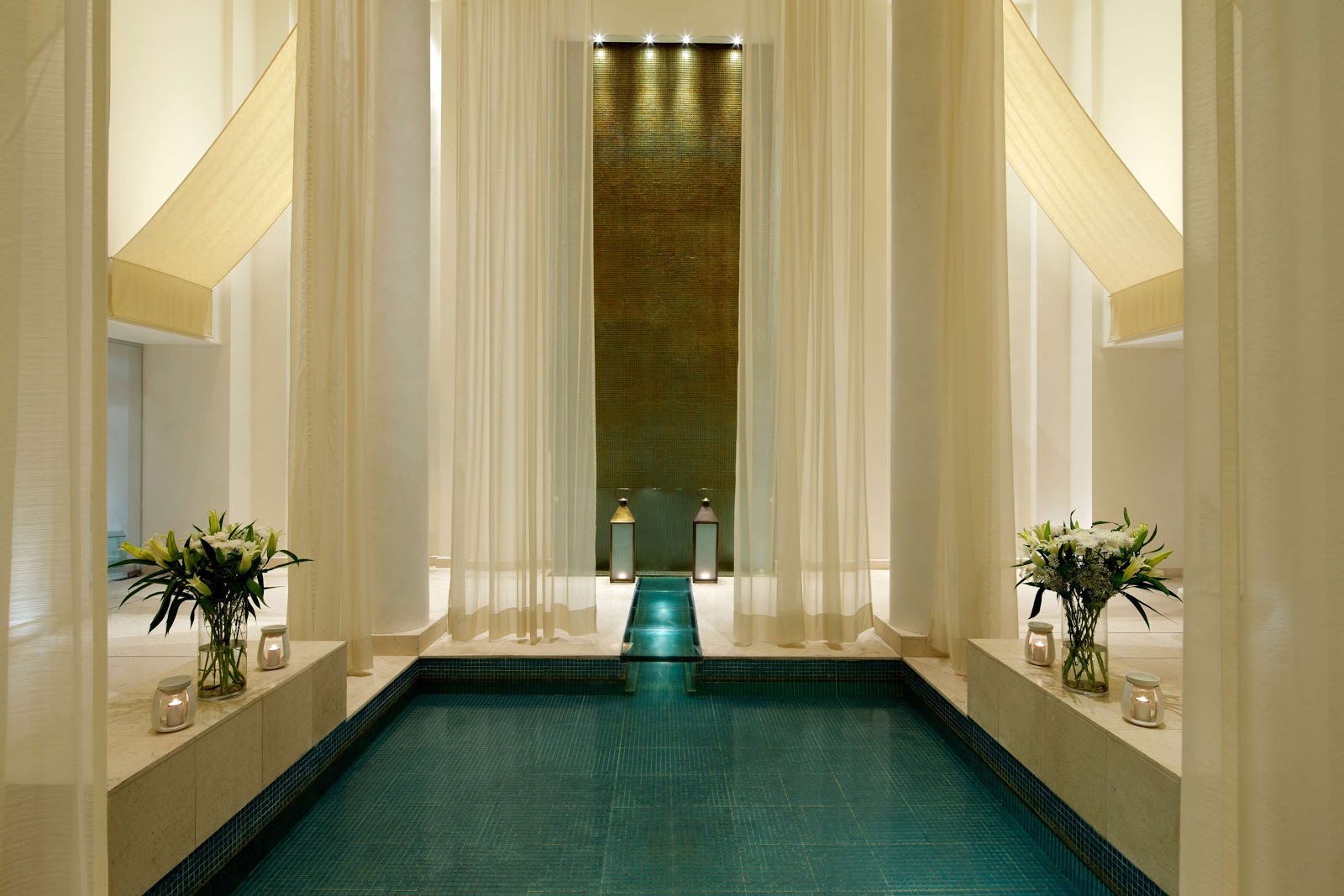 A serene and secluded indoor plunge pool or vitality pool, likely part of a spa, featuring elegant sheer drapes, a waterfall wall, and ambient lighting for a tranquil experience.