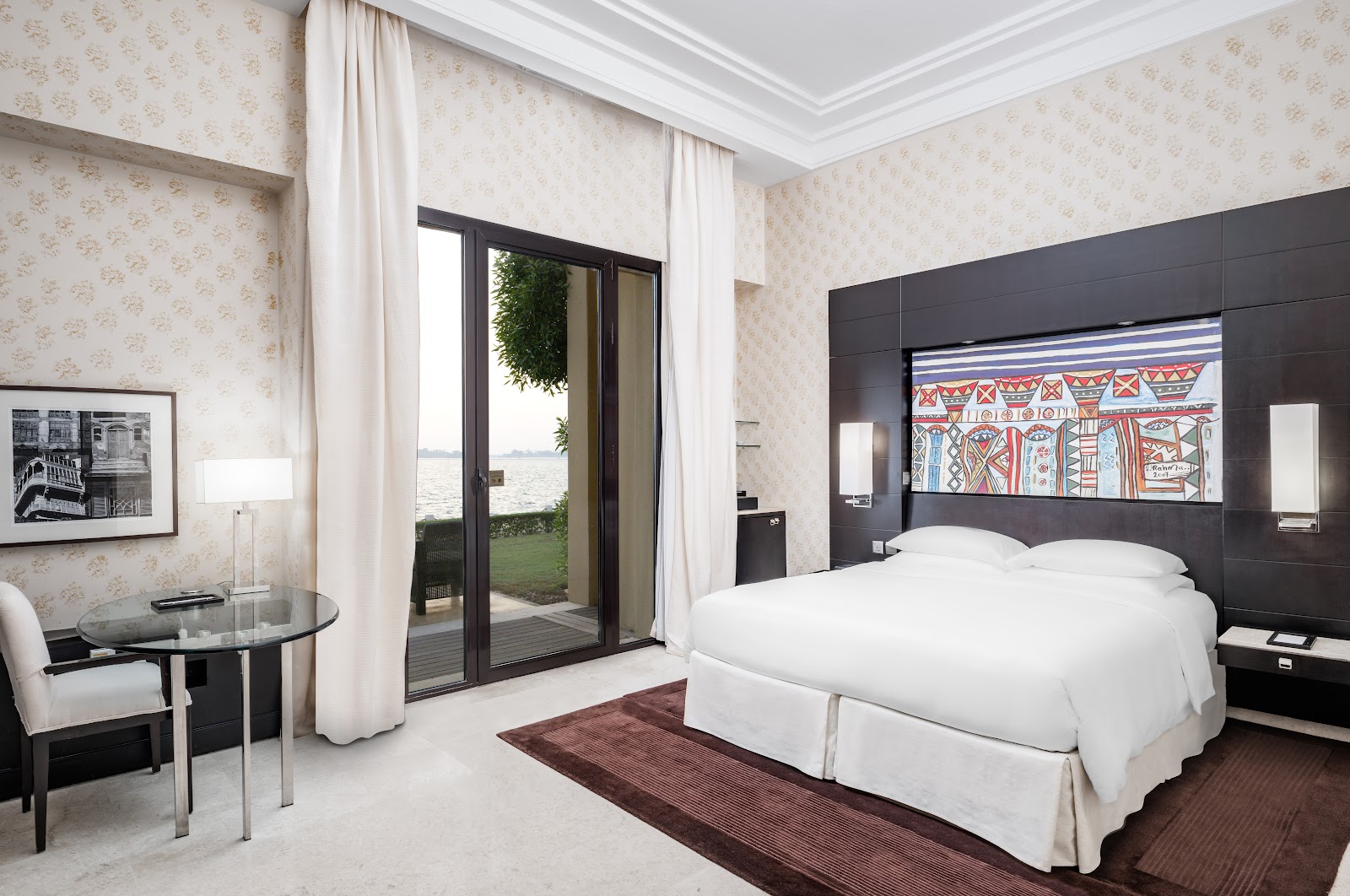 A spacious luxury hotel room with a king-size bed, featuring a private patio or garden access directly overlooking the sea/marina. The room is decorated with modern Arabesque patterns and vibrant local artwork.