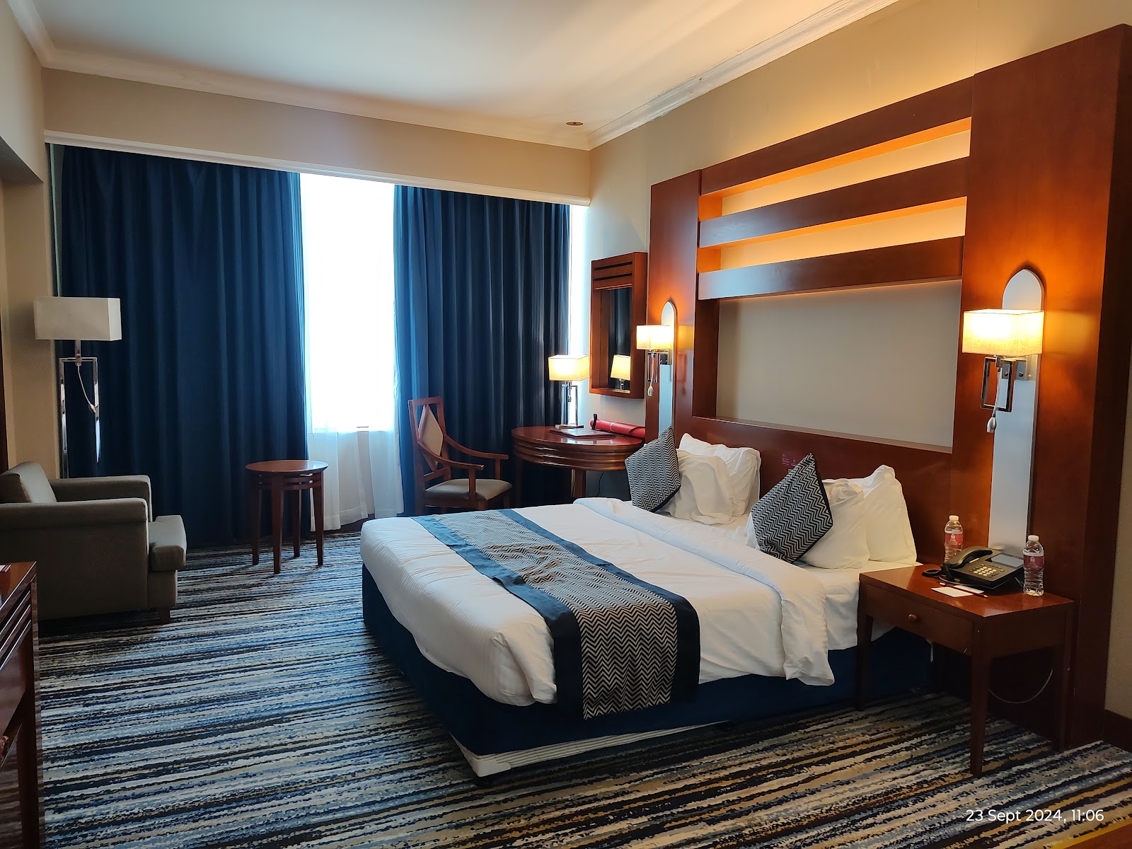 A spacious hotel room featuring a king-sized bed, a comfortable armchair, a work desk, and dark blue curtains.