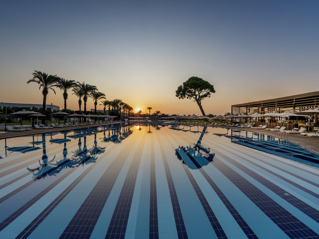 The expansive outdoor pool, identical to image 2, photographed at sunset, reflecting the vibrant colors of the sky. Lined with palm trees and sun loungers.