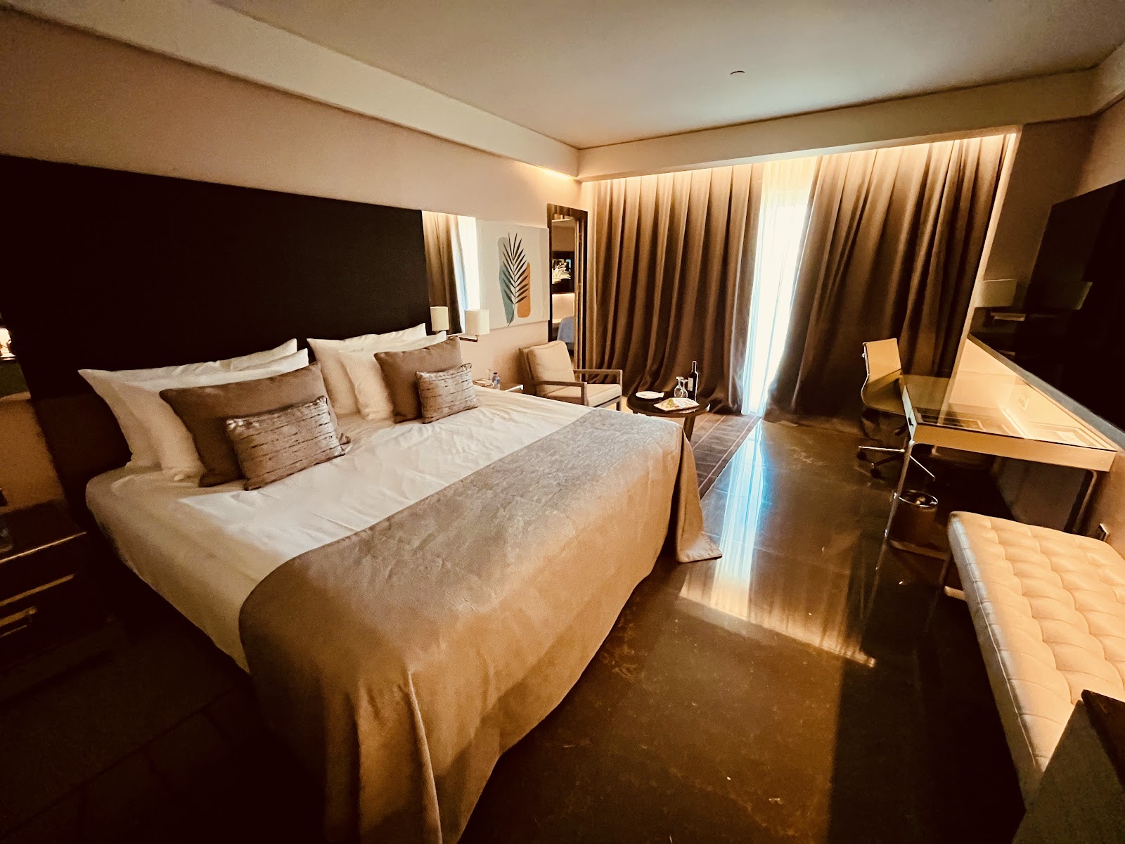 A spacious hotel room with a large king-size bed, dark brown curtains, a desk, and a seating area, featuring a modern, subdued color palette.
