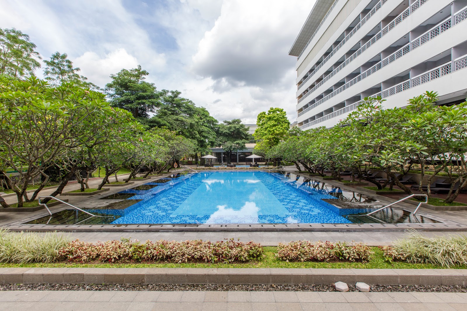 A large, inviting outdoor swimming pool surrounded by lush green trees, part of the Royal Ambarrukmo Yogyakarta's recreational facilities.